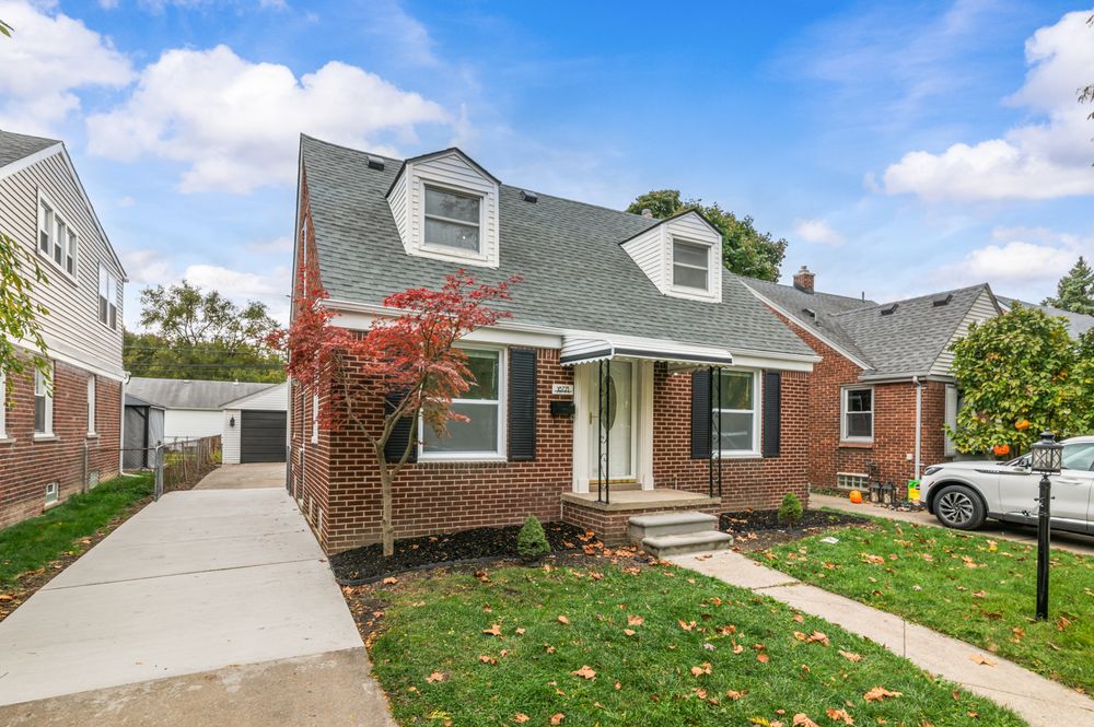 Charming brick Cape Cod home with dormer windows, red maple tree, attached garage, and well-manicured lawn.