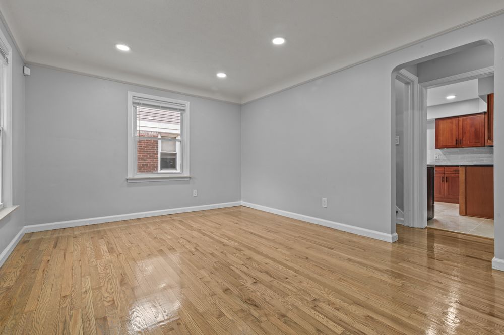 Bright empty living room with gleaming hardwood floors, recessed lighting, crown molding, and an arched doorway leading to a kitchen.