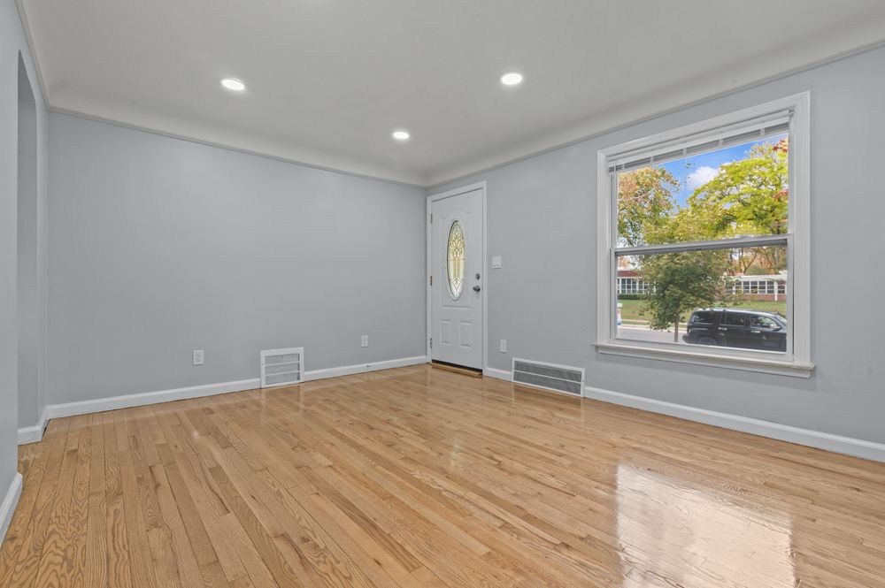 Bright empty living room with hardwood floors, gray walls, crown molding, recessed lighting, and a window with natural light.