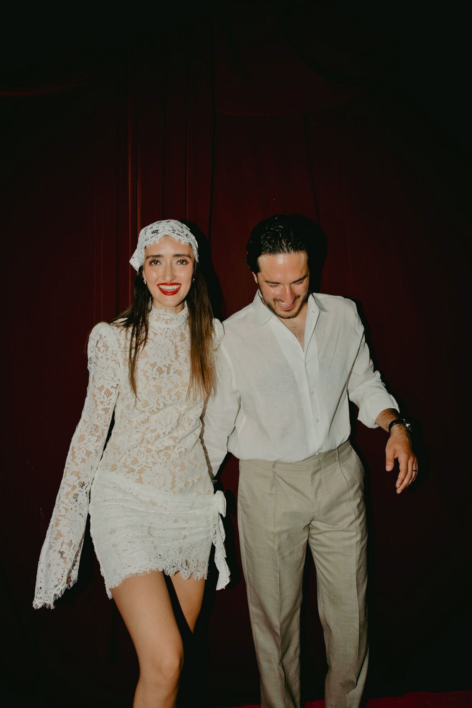 Couple in white outfits dancing together, woman in lace mini dress with headpiece, man in white shirt and beige trousers.