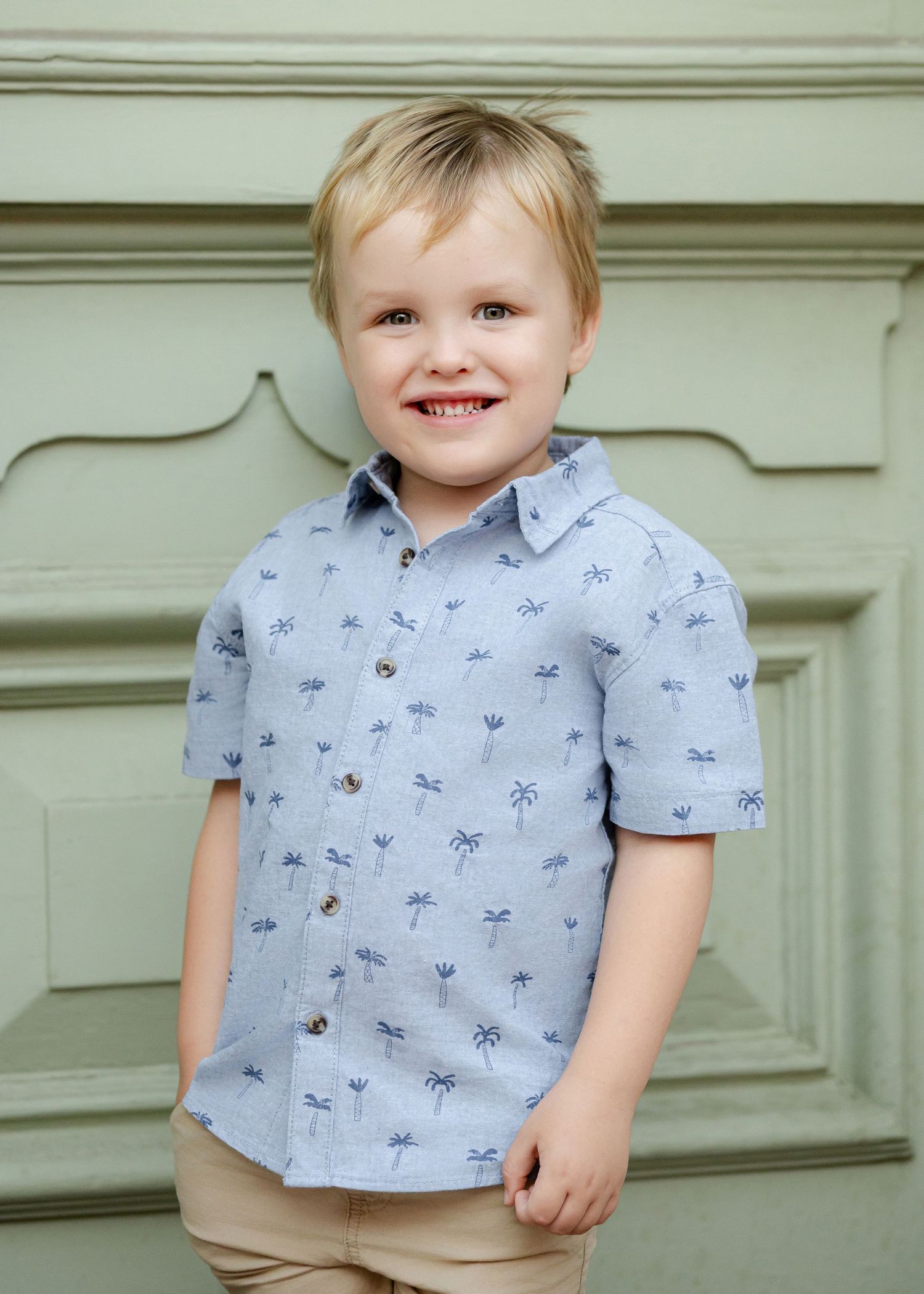 Smiling toddler boy wearing a light blue palm tree print short-sleeve button-up shirt, standing against a sage green door.