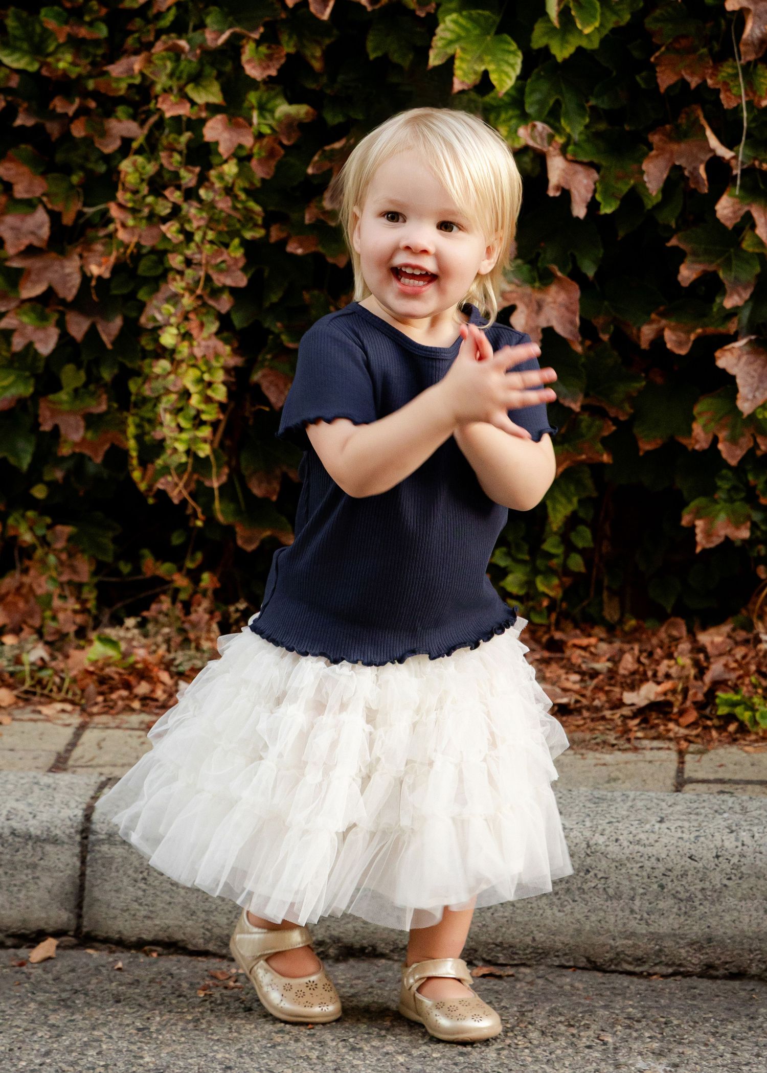 Toddler girl in navy top and white tutu skirt clapping hands, wearing gold shoes, standing outdoors near ivy wall.