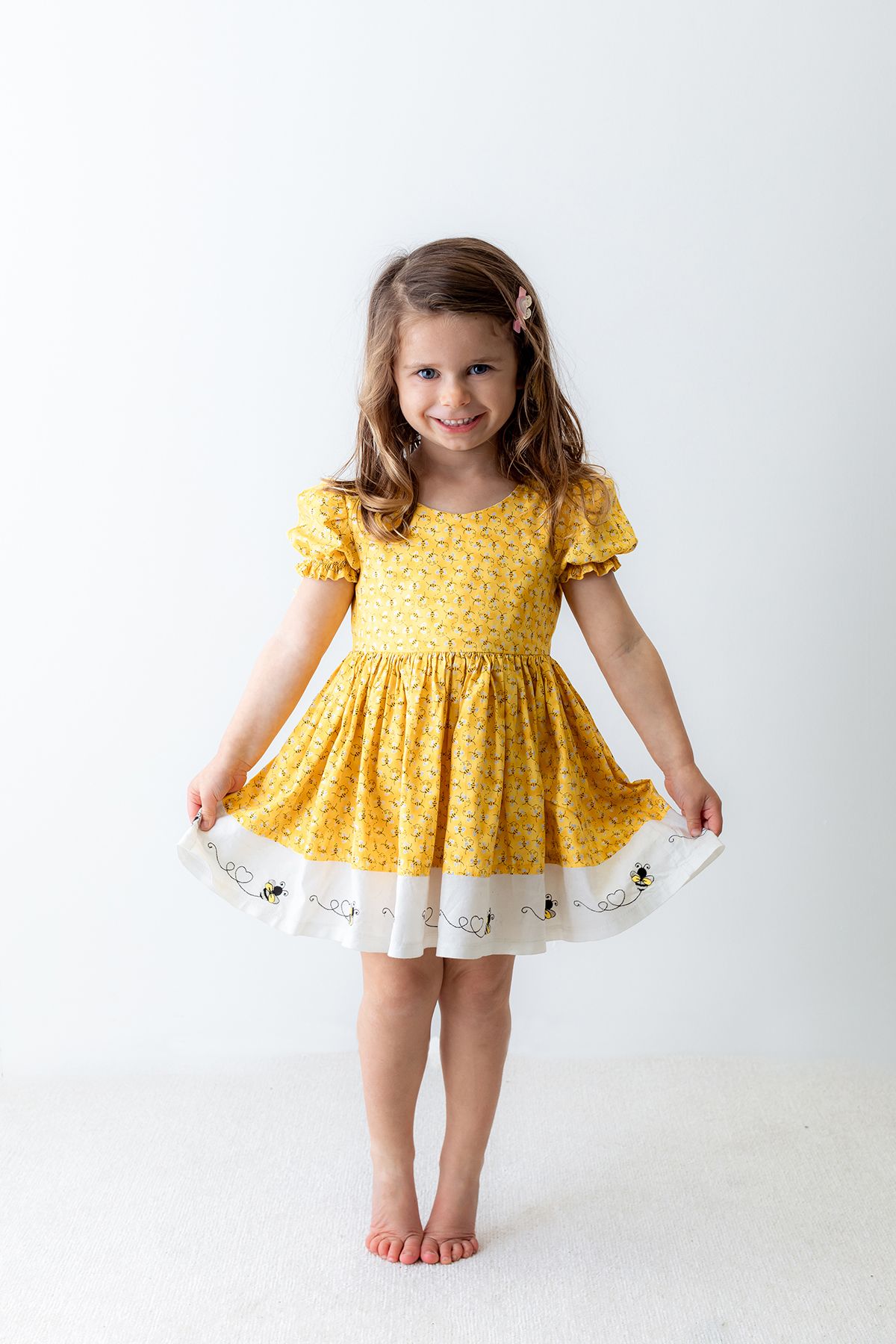 Young girl in yellow floral dress with bee trim, smiling and holding skirt out, standing barefoot on white background.