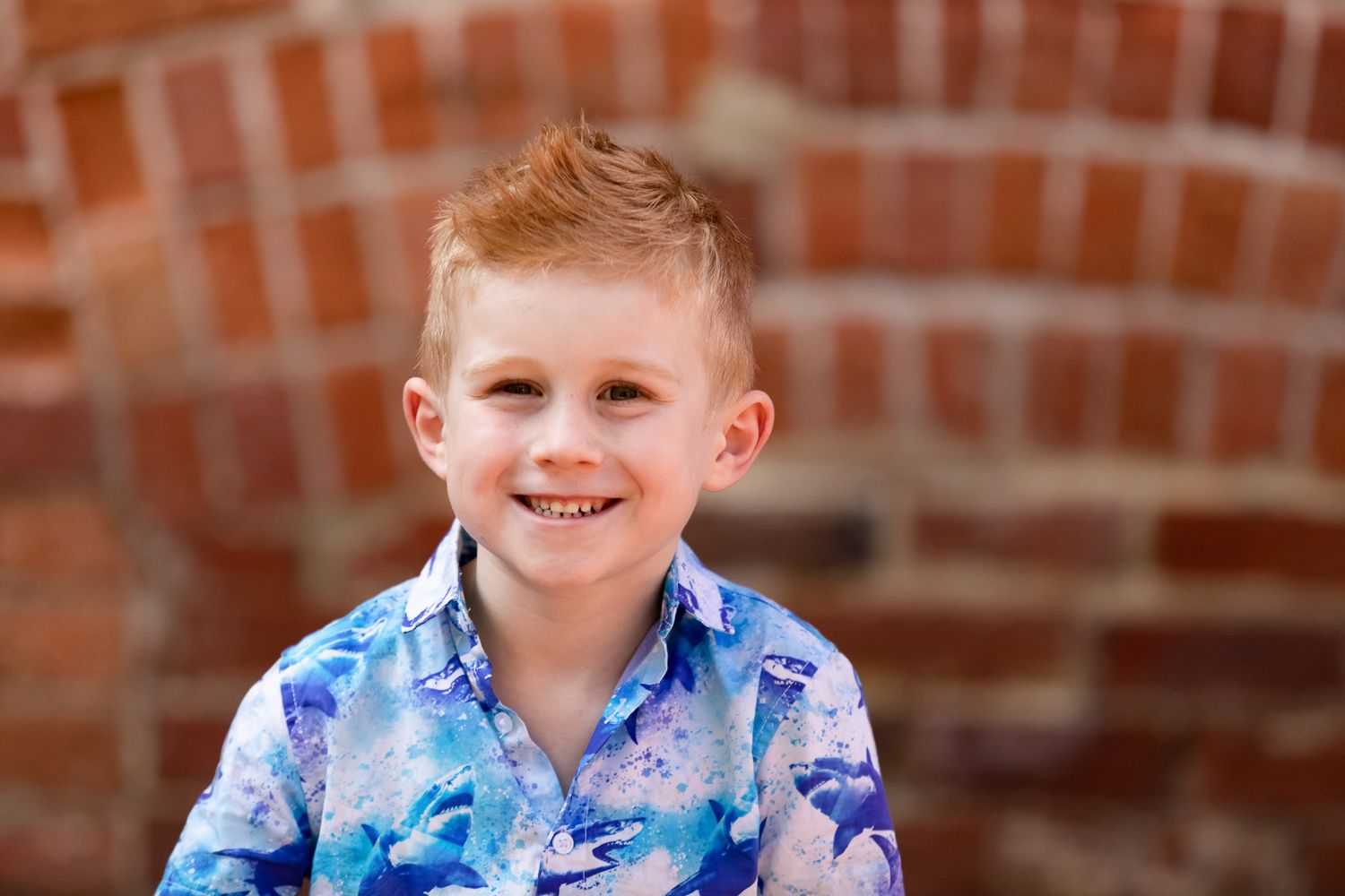 Smiling red-haired boy in a blue floral shirt posing in front of a brick wall backdrop.