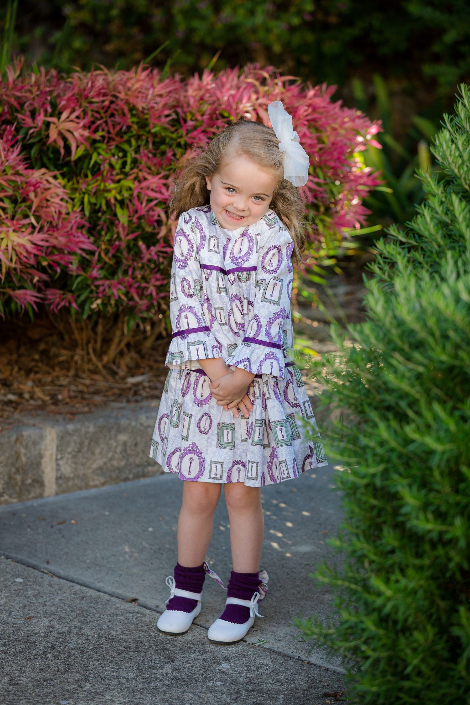 Young girl in purple print dress with bow, posing on garden path surrounded by colorful shrubs.
