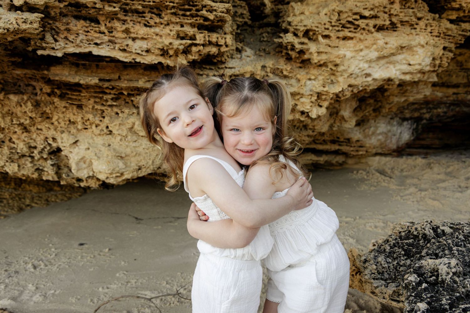 Two young girls in white dresses hugging and smiling on a sandy beach with rocky cliffs behind them.