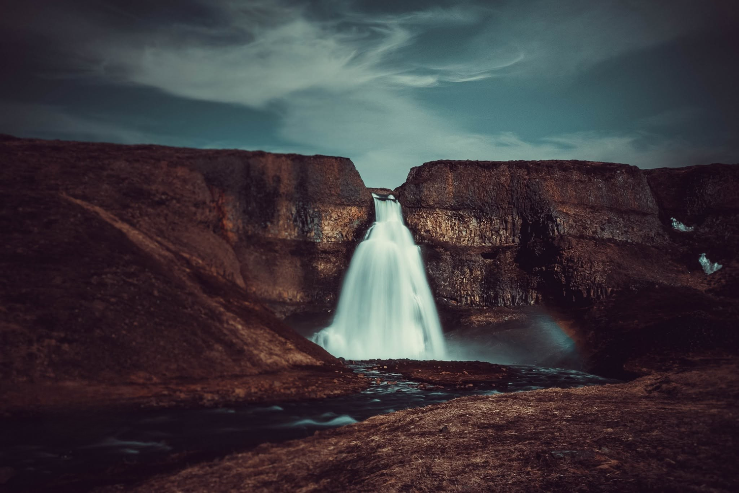 A dramatic waterfall cascades through rugged rocky cliffs under a moody dark sky in Iceland.