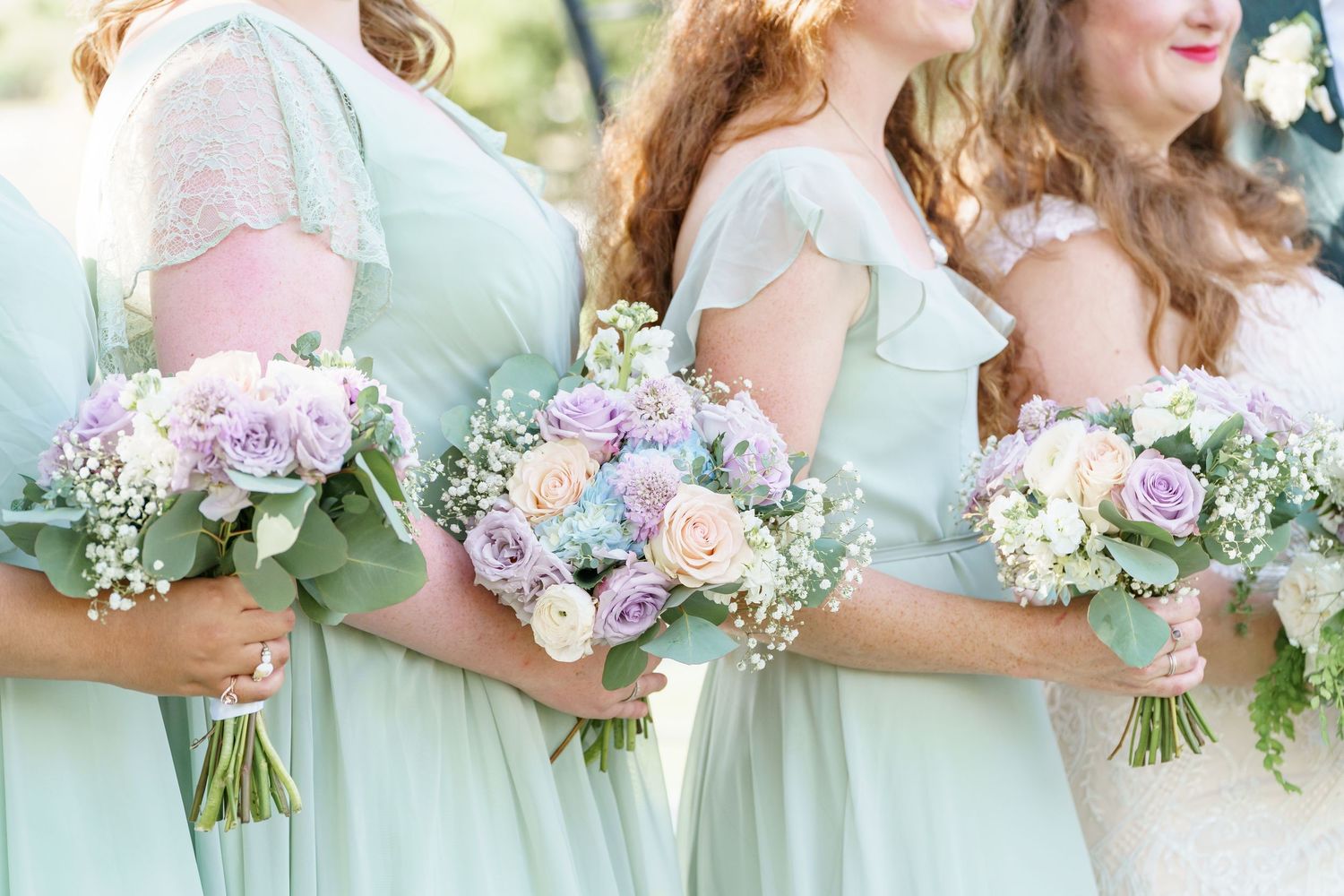 Bridesmaids in mint green dresses holding pastel bouquets of purple, pink, and white flowers at a wedding.