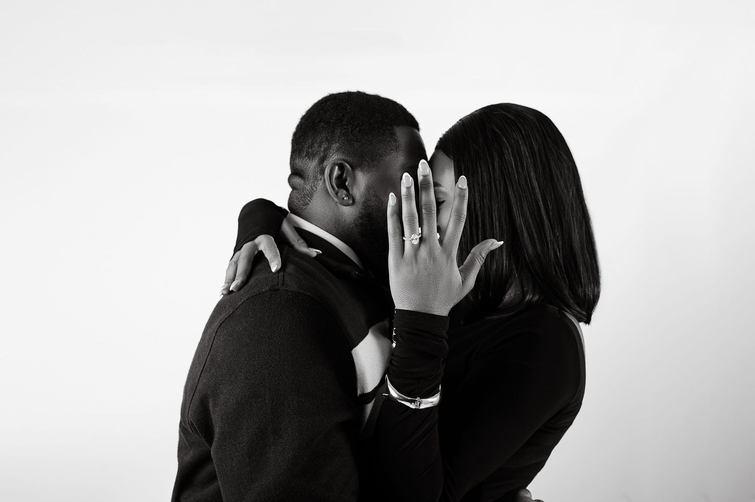 Black and white photo of a couple sharing a kiss, woman showing off engagement ring, romantic proposal moment.