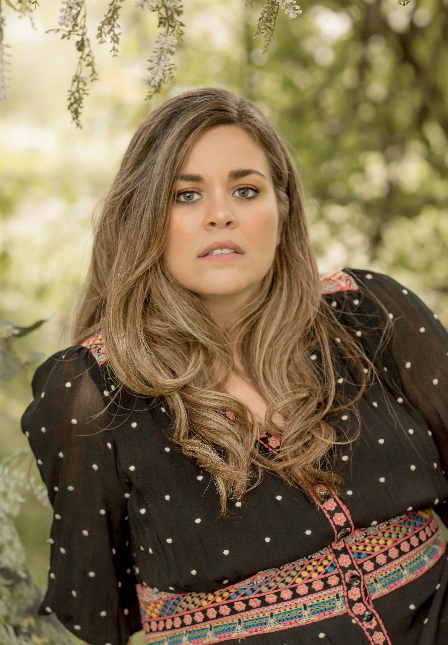 Brunette woman in a black floral dress posing outdoors among blooming trees.