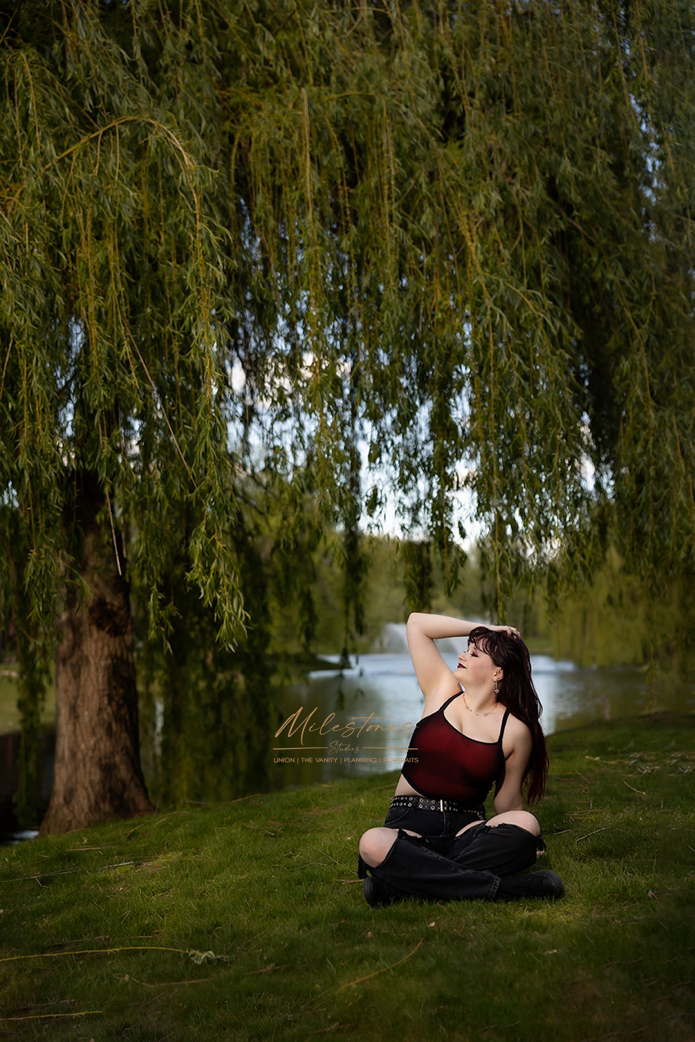 Senior girl posed under a weeping willow tree wearing jeans and a red top in Eagle Idaho. Milestones Studios LLC