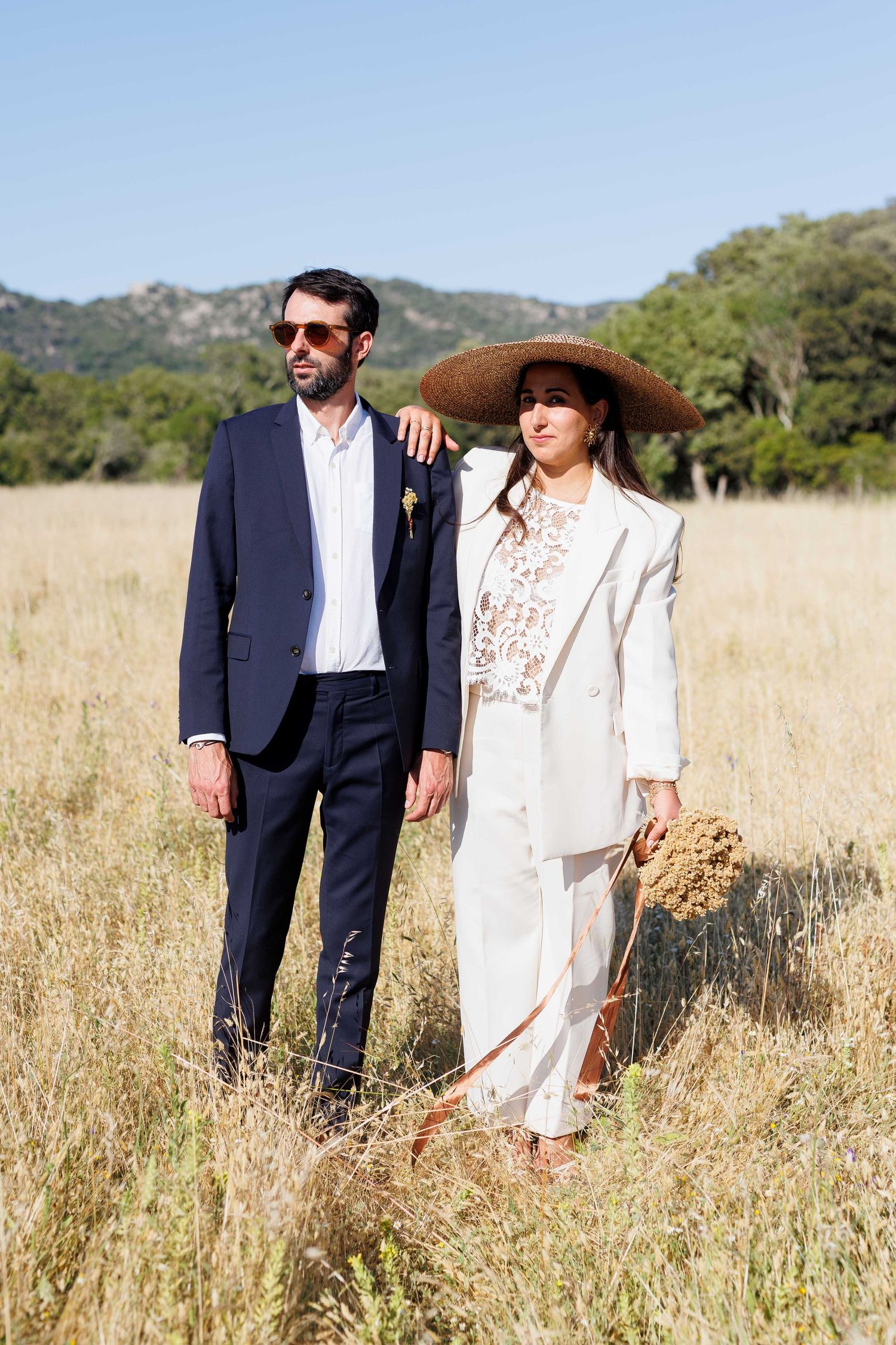 Couple in a golden field, man in navy suit with sunglasses, woman in white suit and straw hat, mountains behind.
