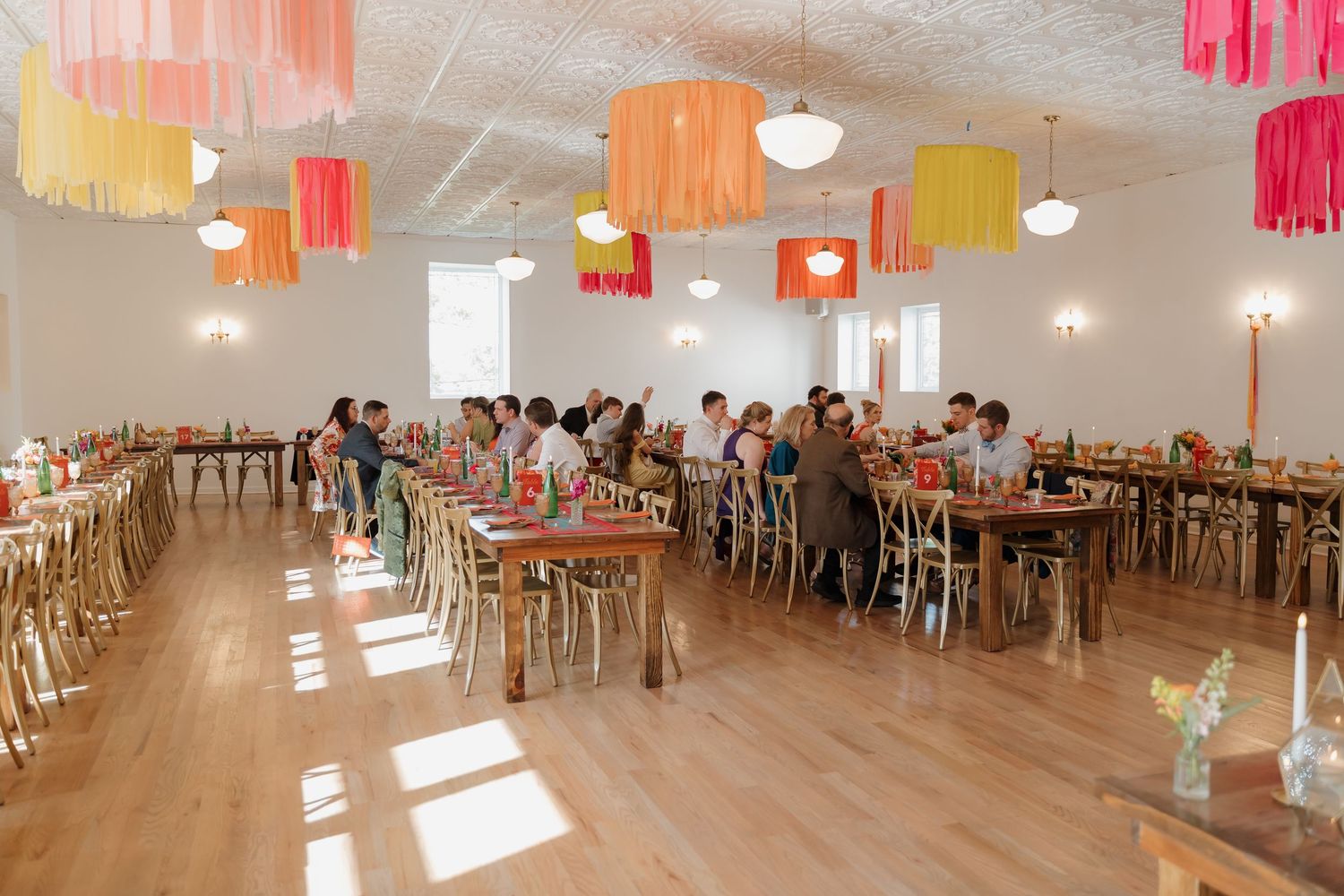 Colorful fringe ceiling decorations hang above long wooden dining tables filled with guests at a bright, modern event venue.