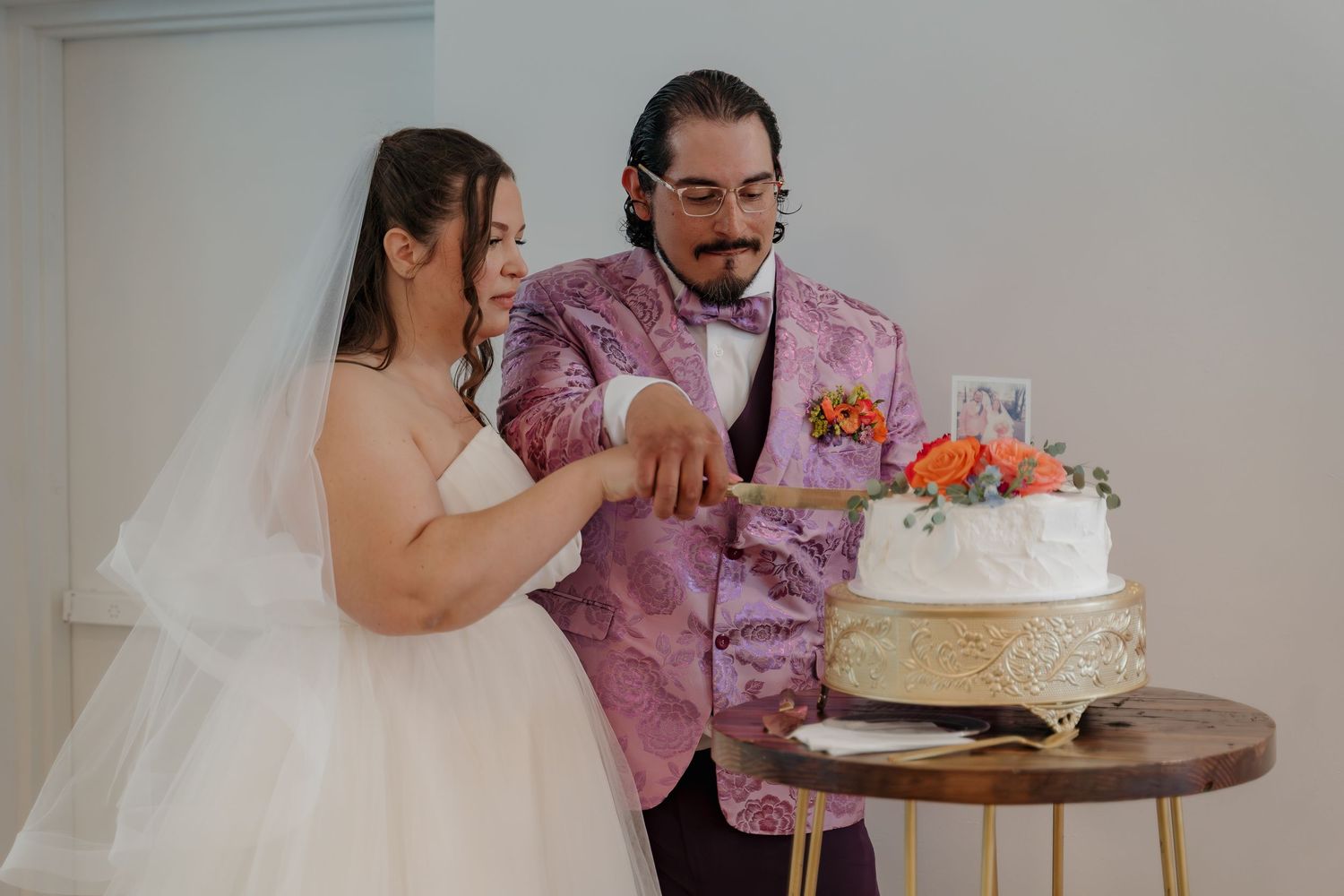 Bride and groom cutting their white wedding cake together, groom wearing a pink floral blazer with bow tie.