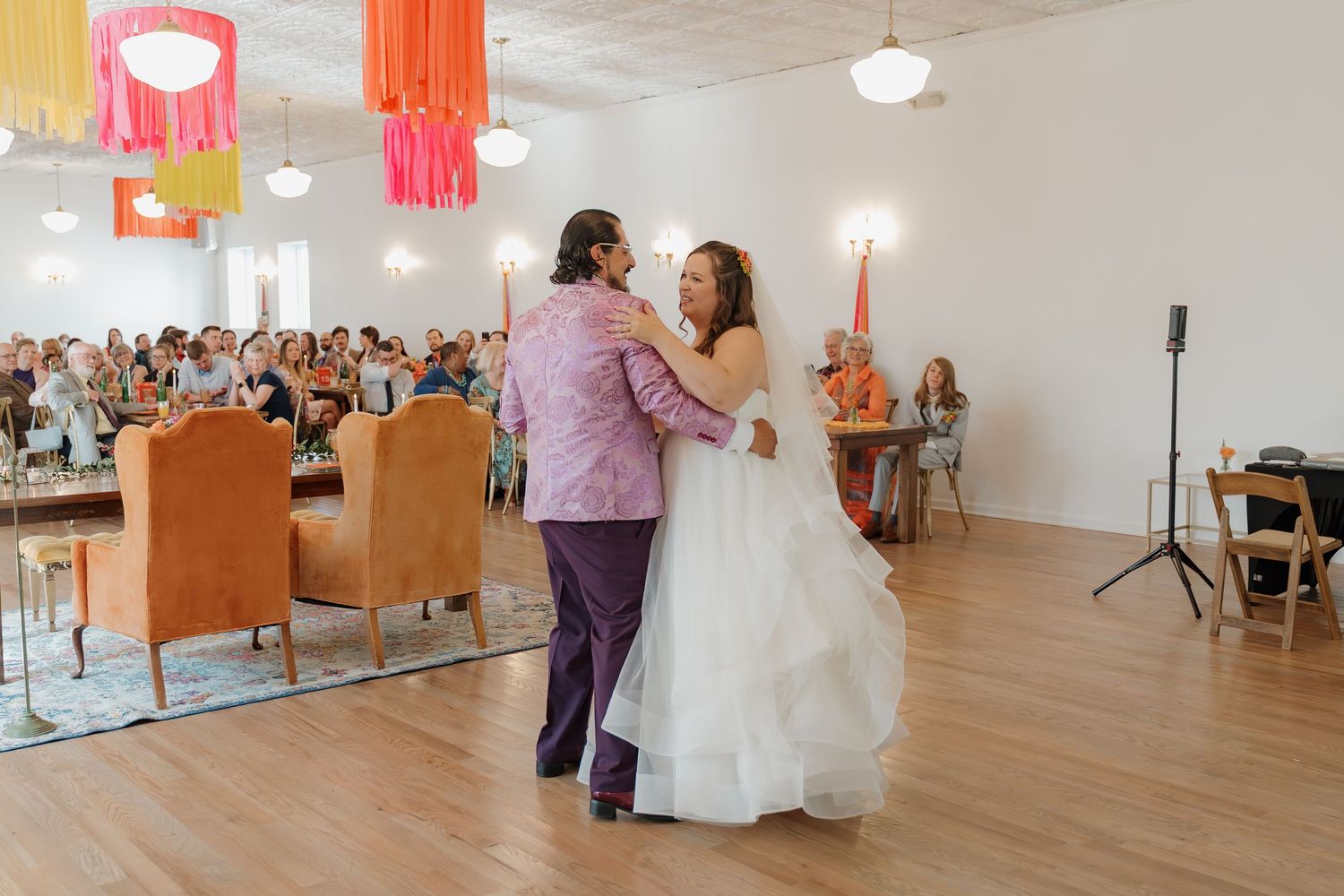 A bride and groom share their first dance in a bright reception hall filled with colorful hanging decorations.