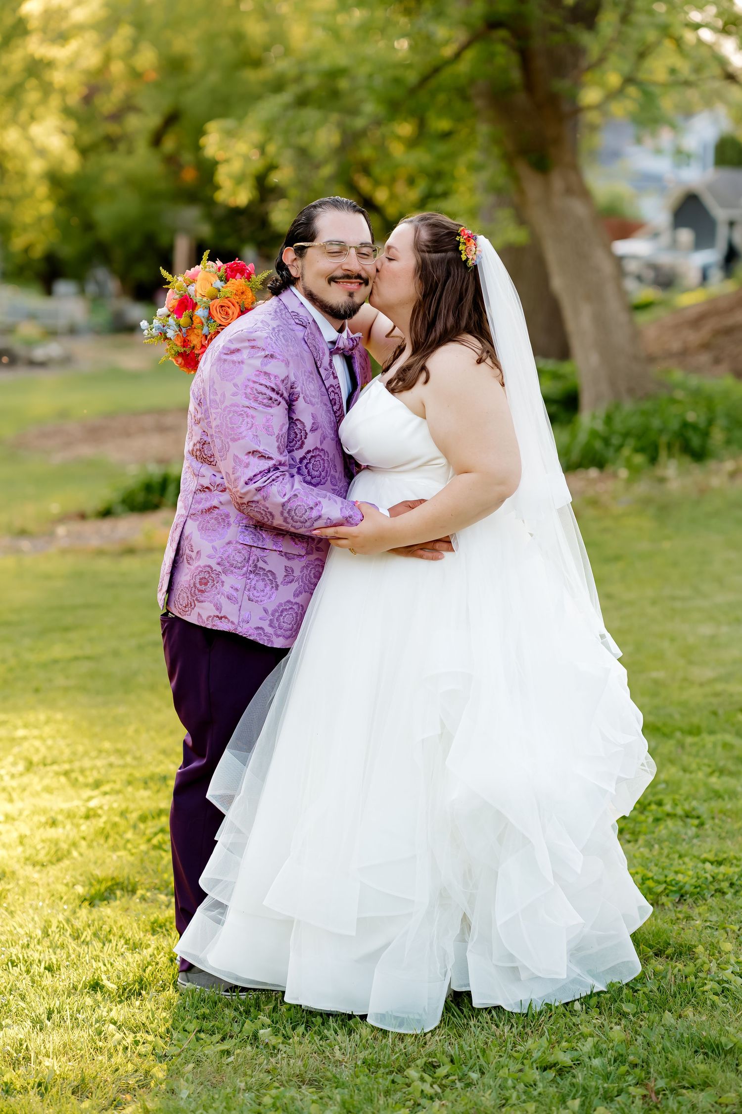 Bride in white ruffled gown kisses groom in purple paisley jacket outdoors on green lawn.