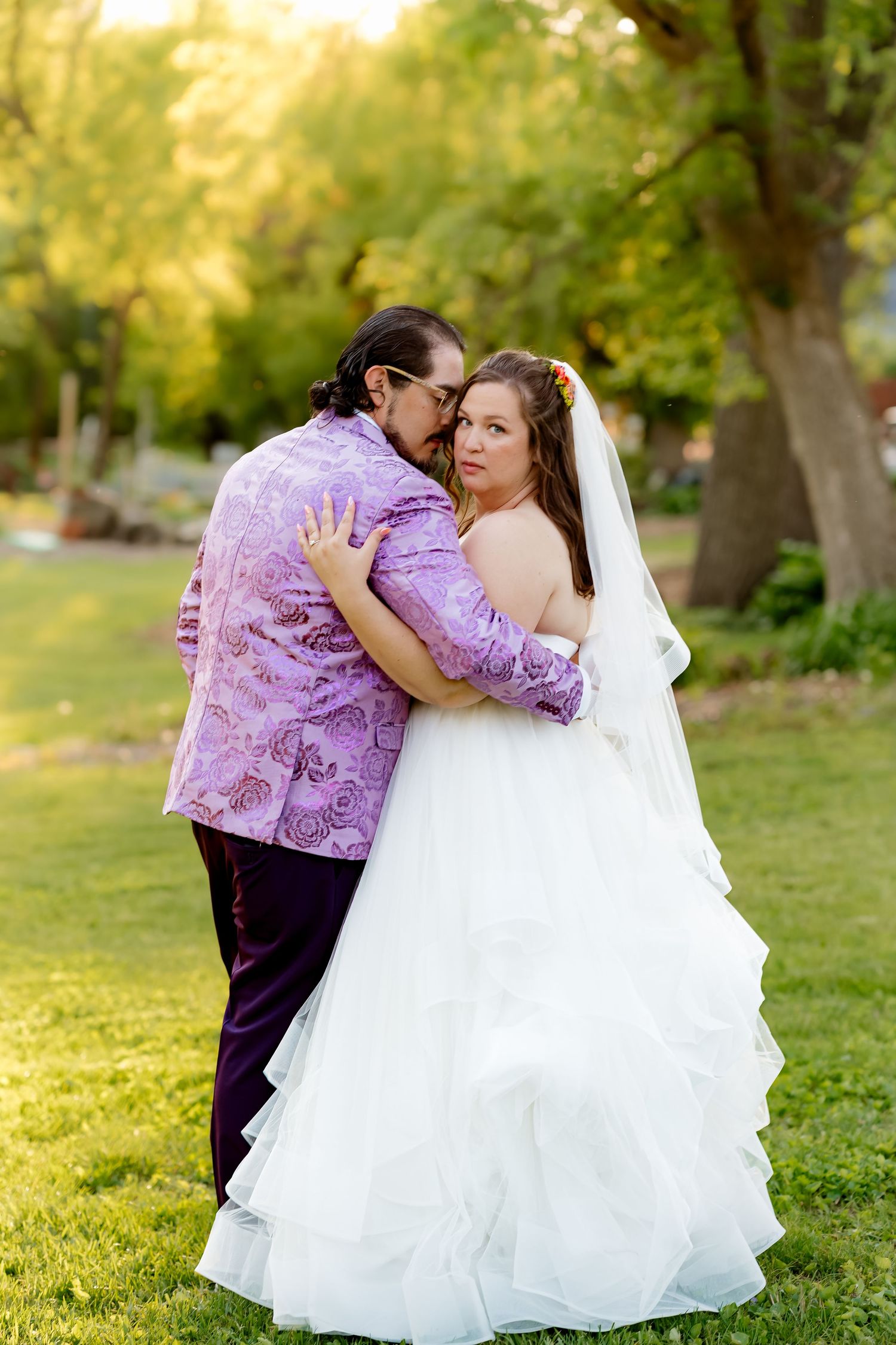 Bride in white ruffled gown and veil embraces groom in purple paisley jacket outdoors in lush green garden setting.