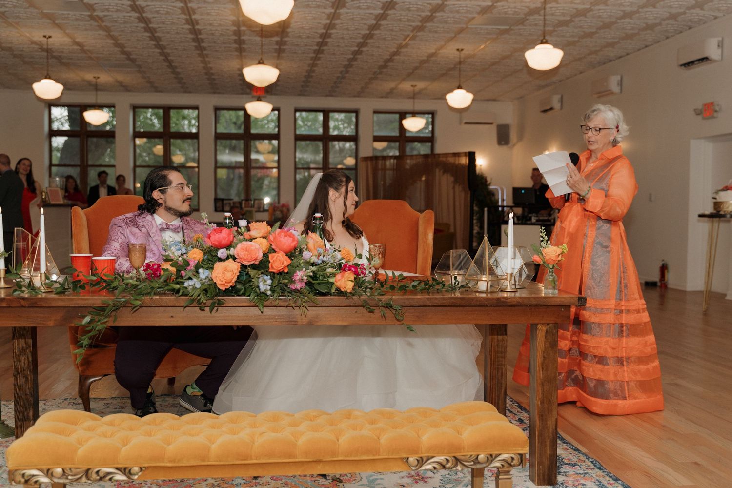 Bride and groom sit at sweetheart table as officiant in orange dress delivers wedding speech indoors.