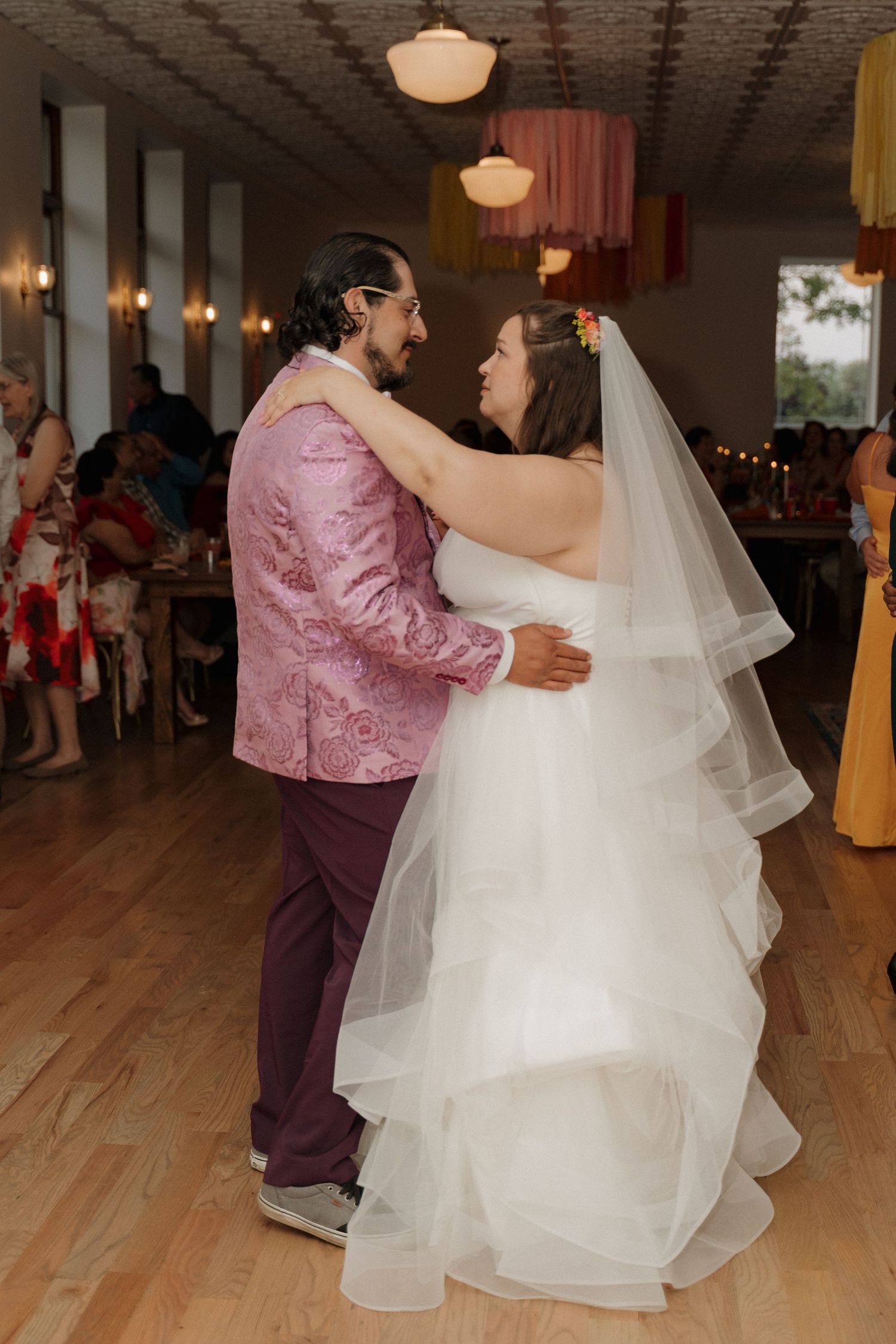 Bride in layered white gown and veil shares first dance with groom in pink paisley jacket on wooden floor.