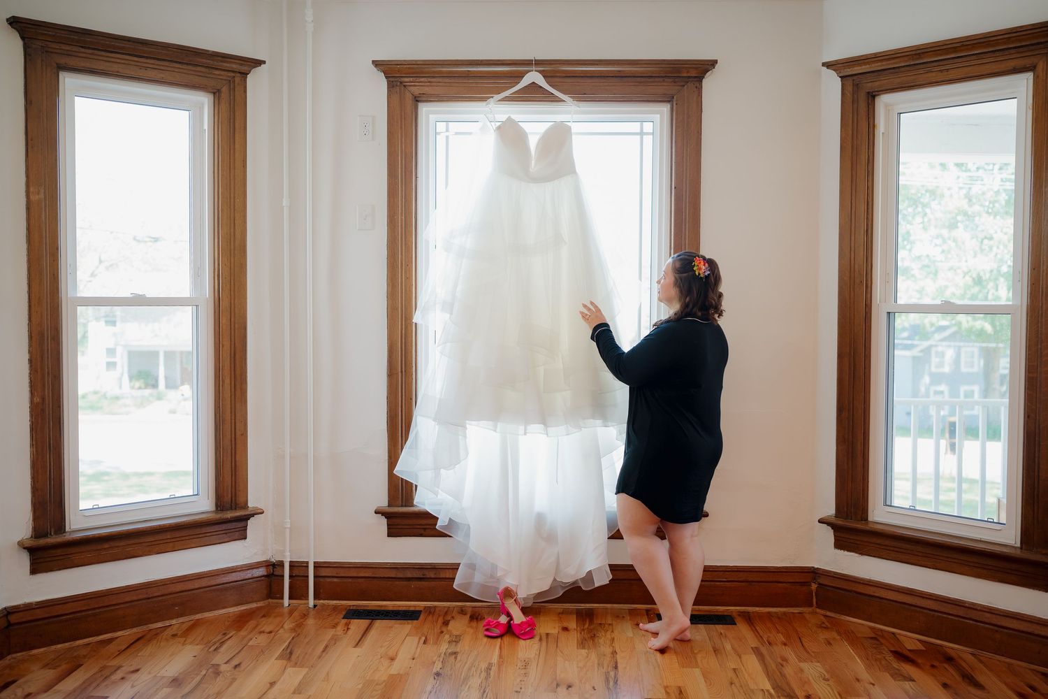 Bride admiring her white wedding gown hanging in a sunlit bay window on hardwood floors.