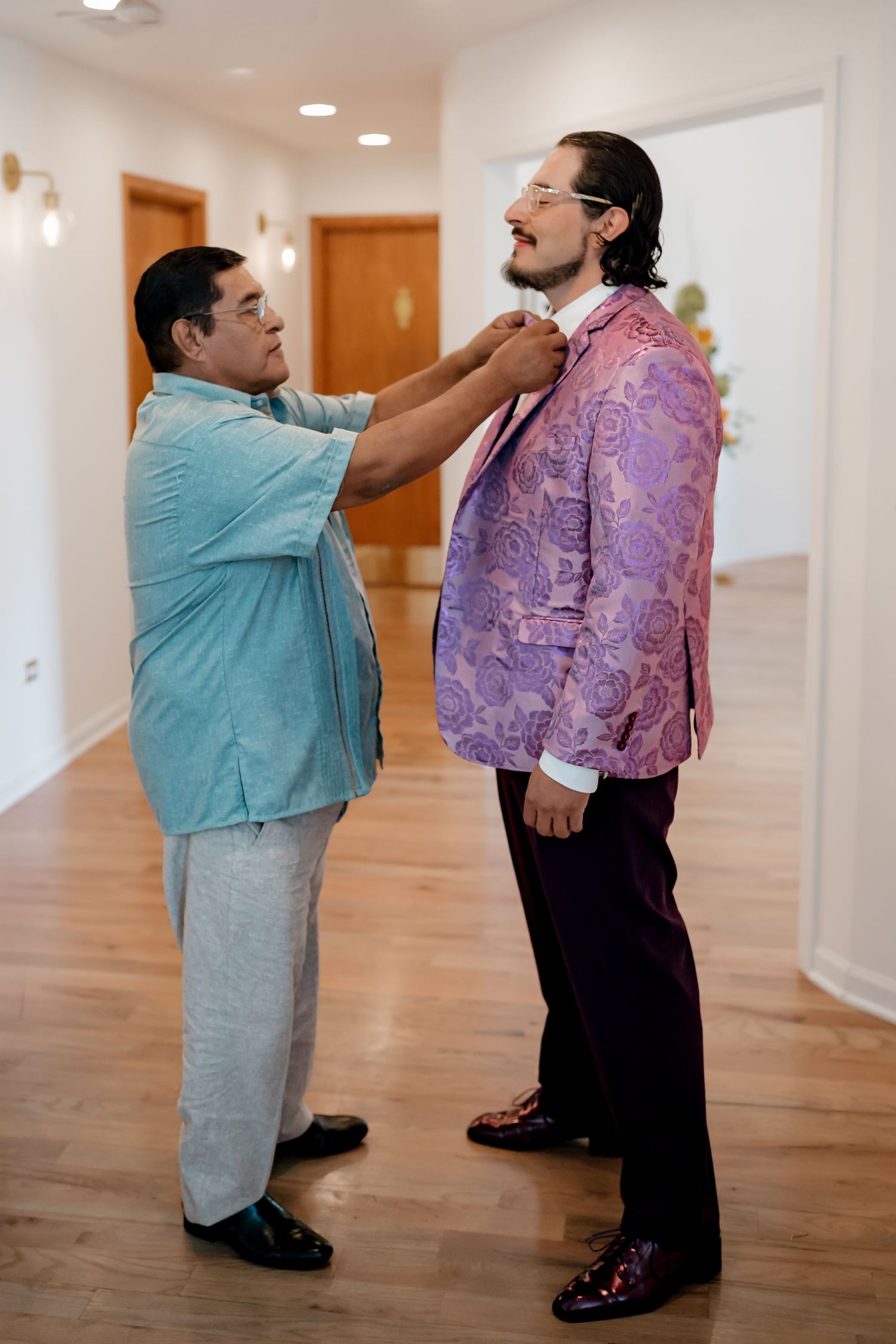 A man in a teal suit adjusts the tie of a groom wearing a purple floral blazer before a wedding ceremony.