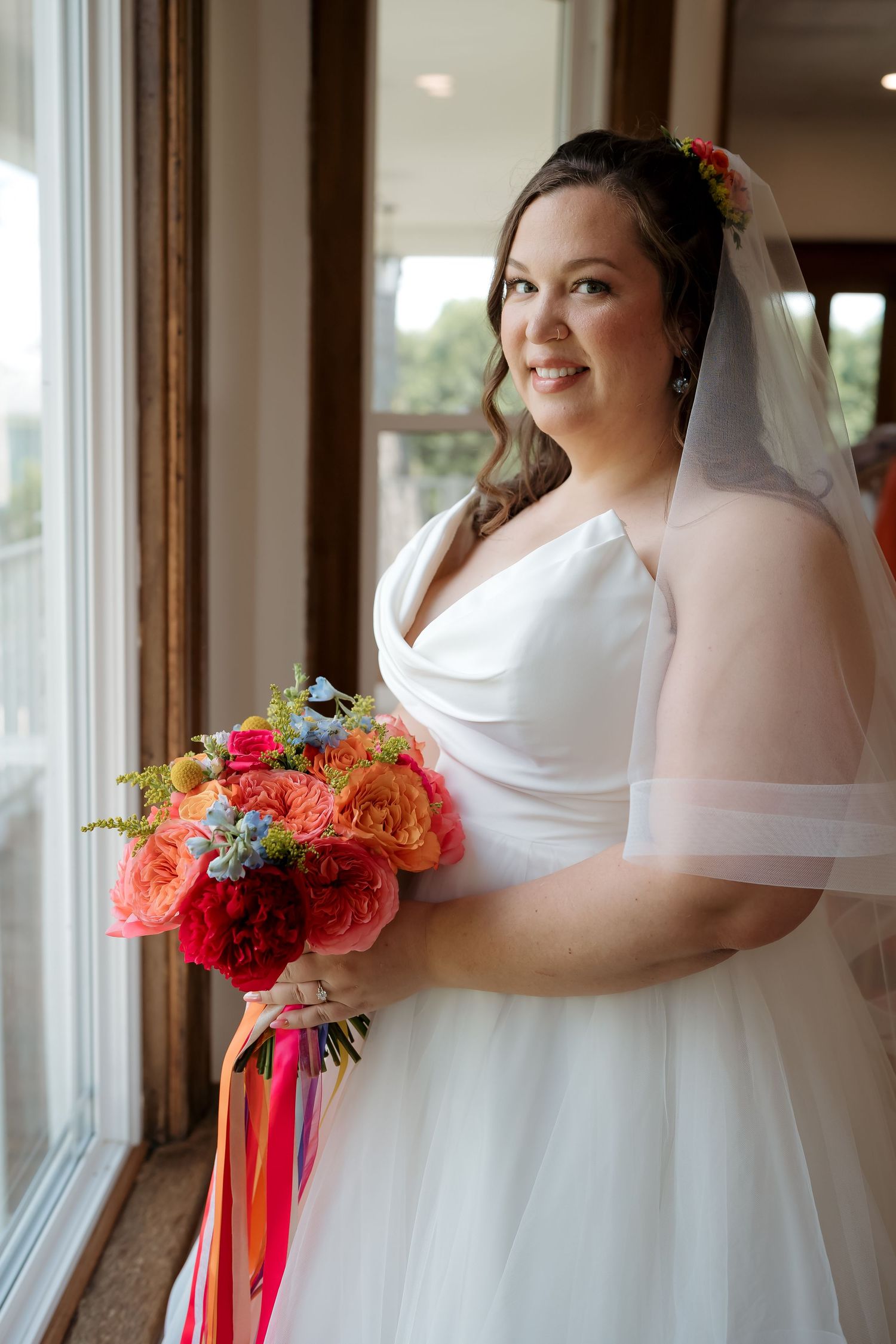 Smiling bride in white gown and veil holds colorful bouquet of red, orange, and blue flowers by a window.