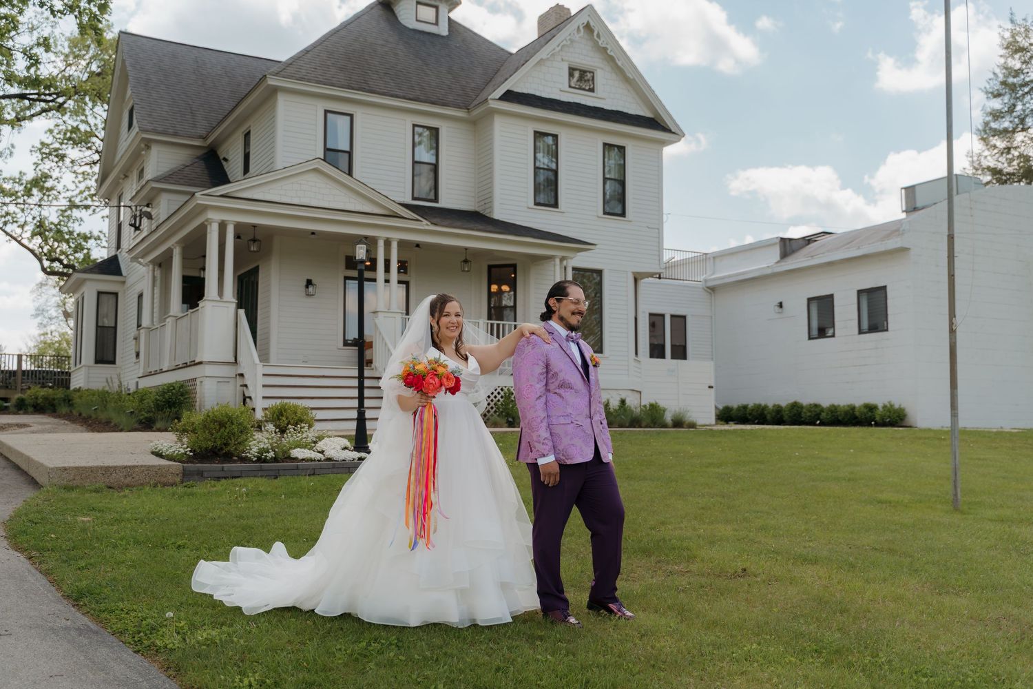 Bride in white ballgown and groom in purple suit pose outside a historic white Victorian wedding venue.
