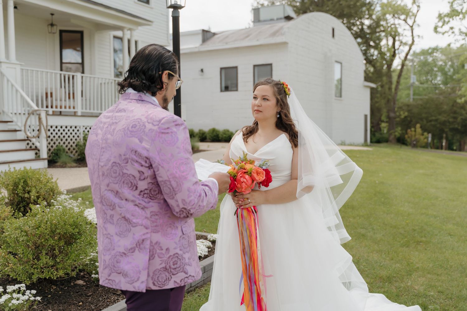 Bride in white gown with colorful bouquet faces groom in purple floral suit during outdoor wedding ceremony.