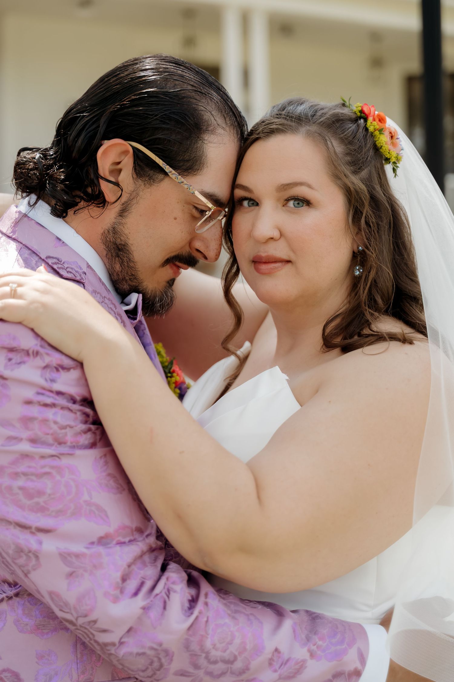 Bride and groom embrace tenderly in wedding attire, bride wearing veil with floral accents, groom in lavender floral suit.