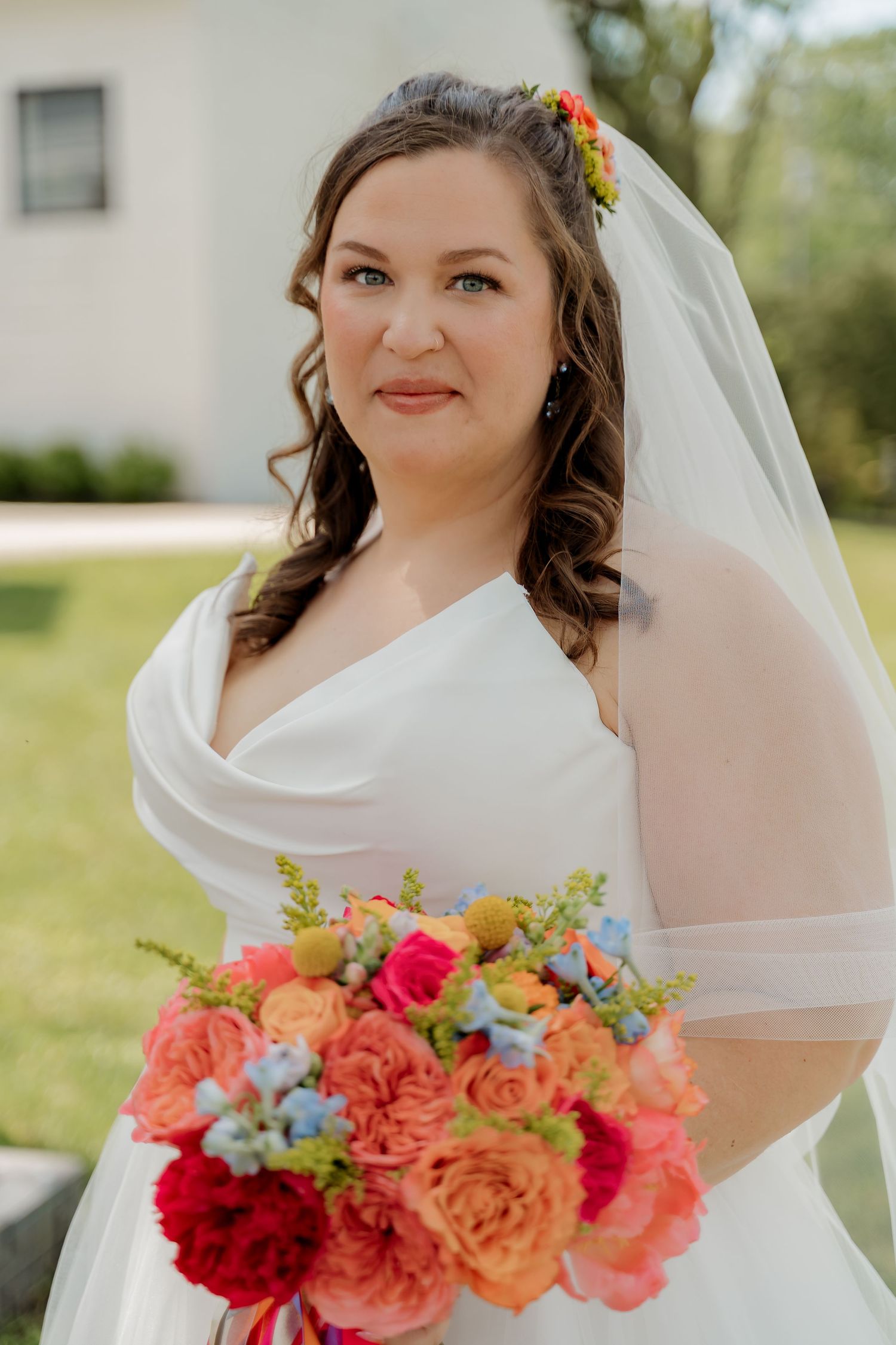 Bride in white wedding dress and veil holds colorful bouquet of pink, orange, and blue flowers outdoors.
