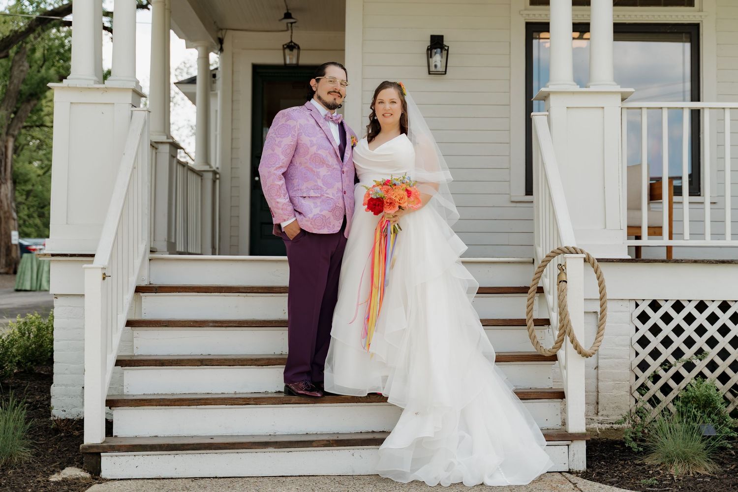 Bride and groom pose on white porch steps, she in white gown, he in pink jacket and purple pants.