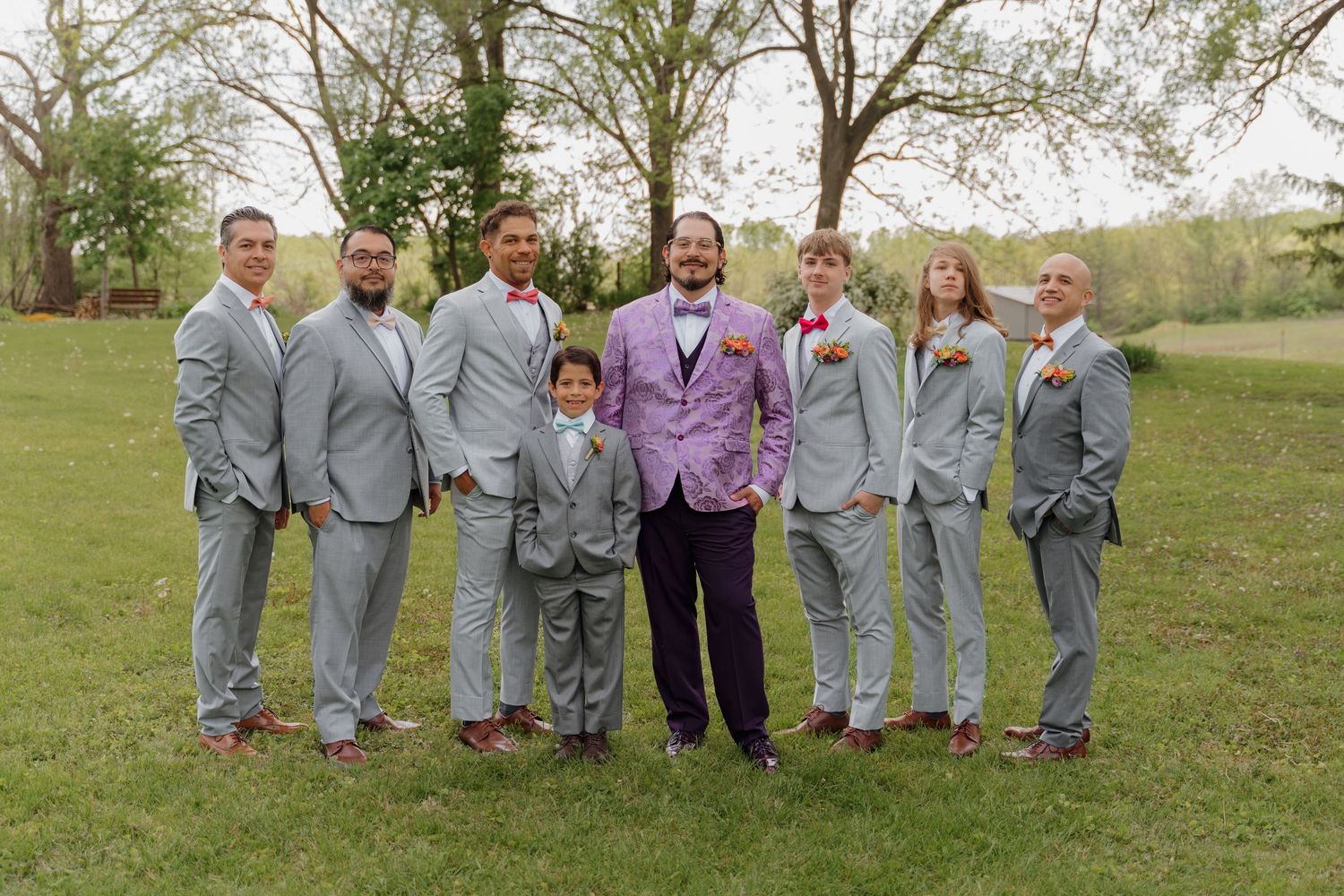 Groom in purple suit stands with groomsmen in gray suits on green lawn during outdoor wedding ceremony.