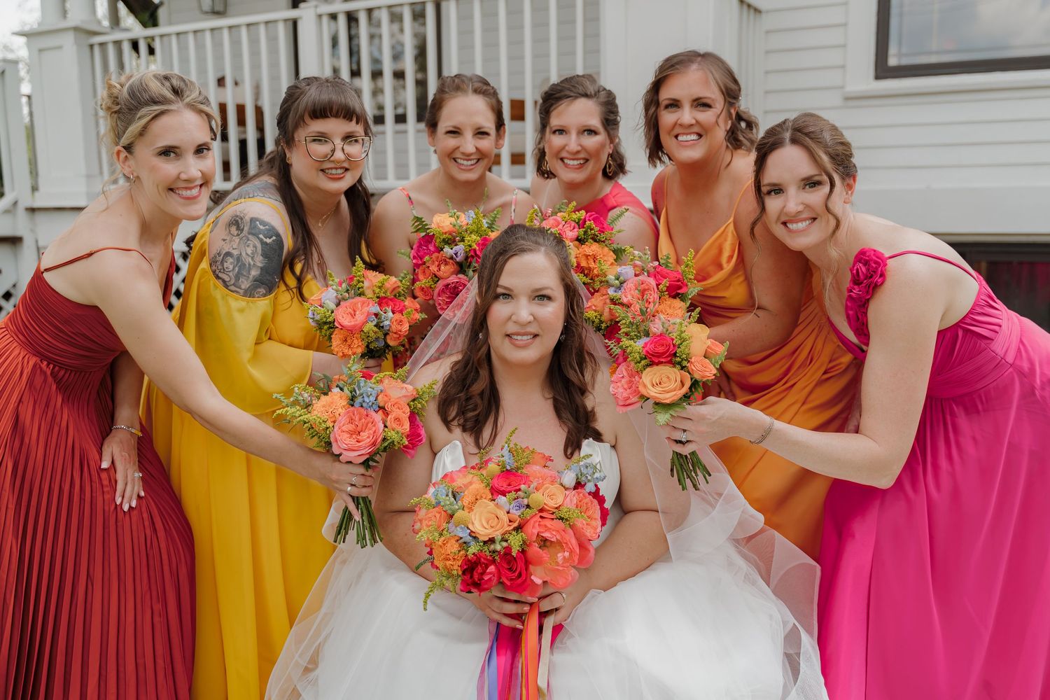 Bride in white dress poses with six bridesmaids in pink, yellow, and orange gowns holding vibrant bouquets.