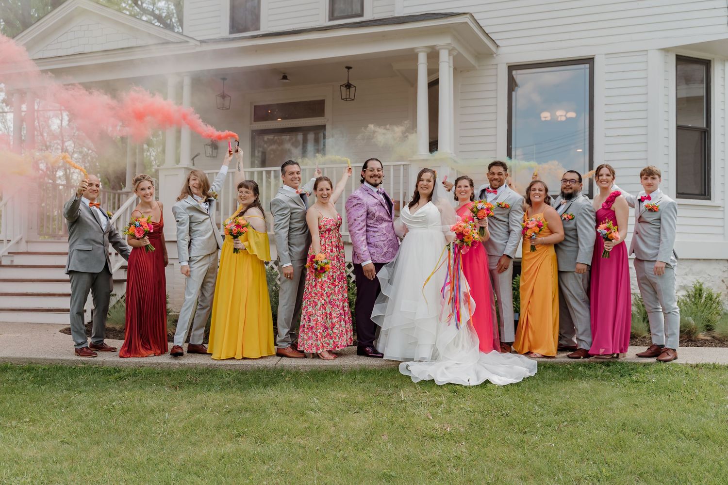 Colorful wedding party poses outside white house with pink smoke bomb, bridesmaids in vibrant yellow, pink, and orange gowns.