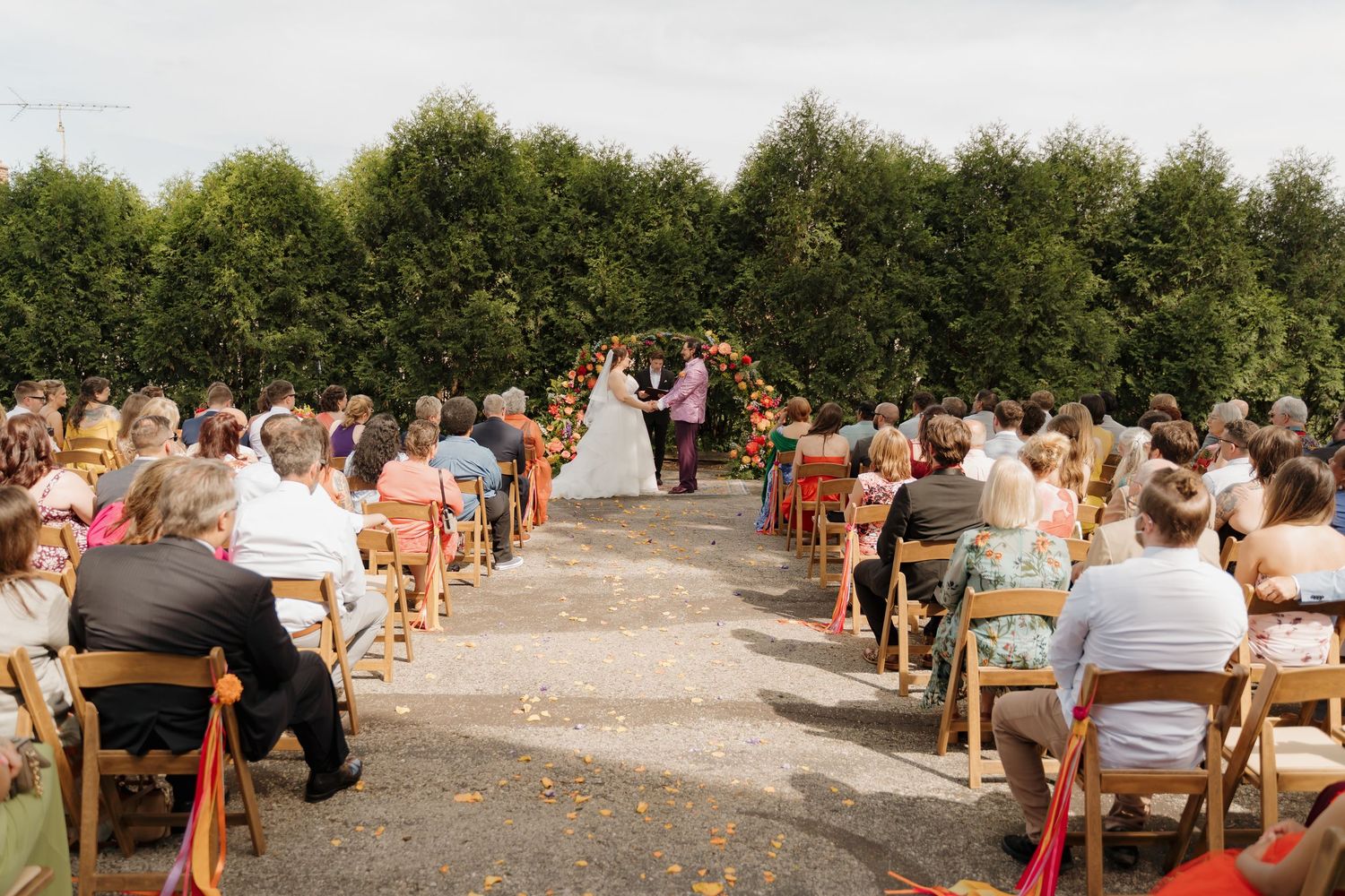 Guests seated in wooden chairs watch a couple exchange vows at an outdoor wedding ceremony with a floral arch.