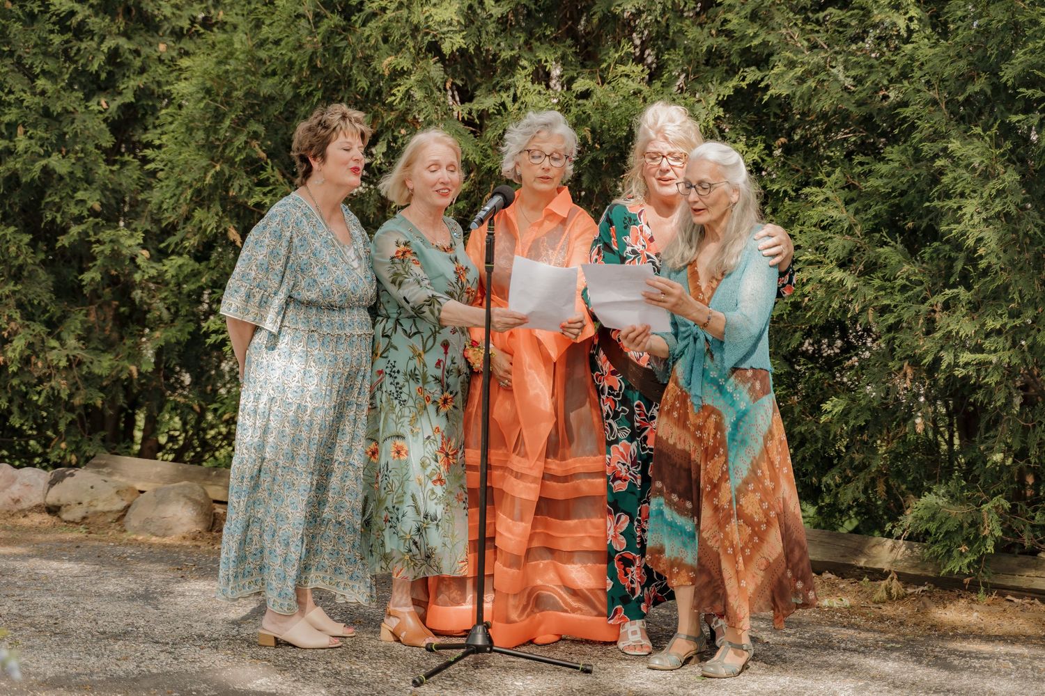 Five women in colorful boho dresses sing together at a microphone stand during an outdoor wedding ceremony.