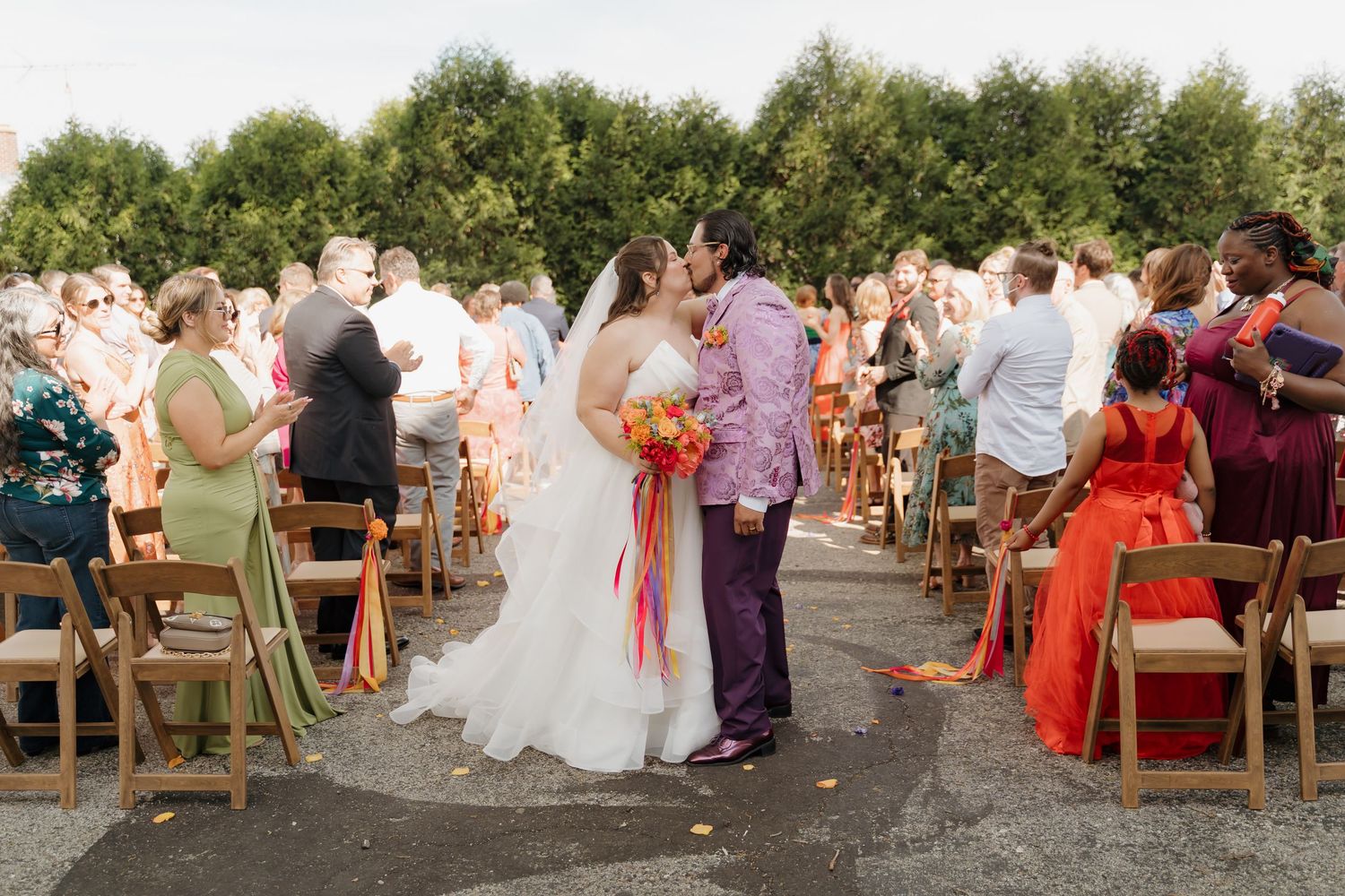 Bride and groom share first kiss at colorful outdoor wedding ceremony with guests in vibrant attire.