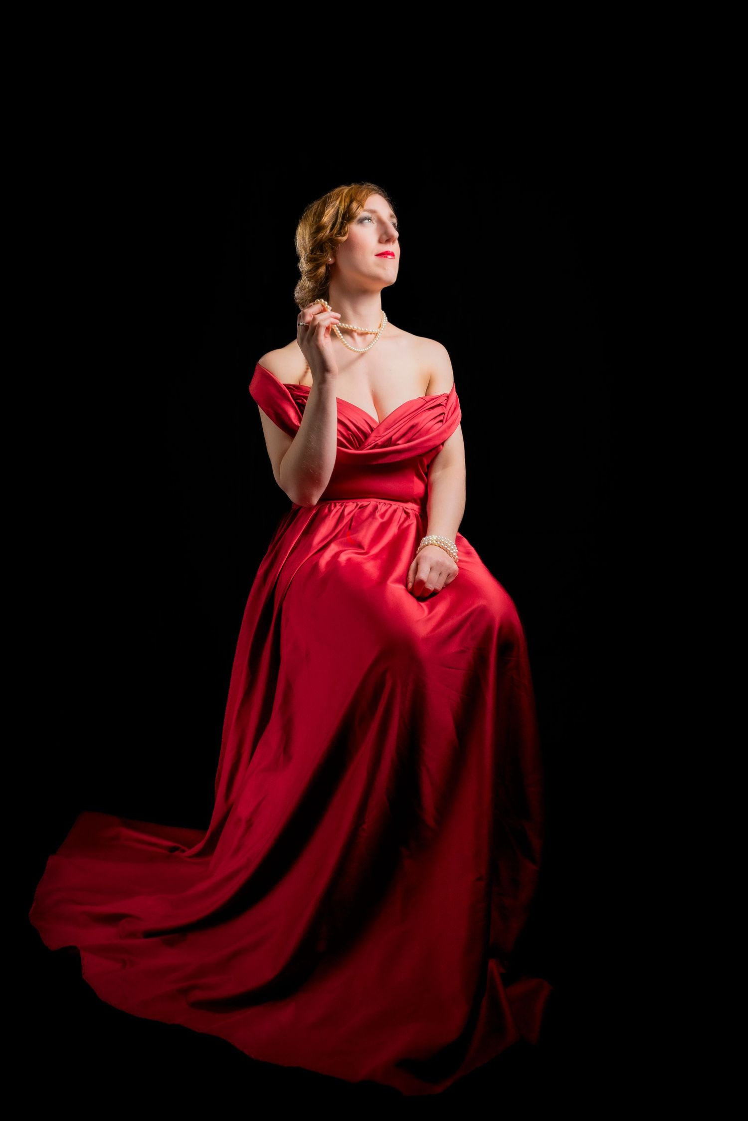 Woman in elegant red off-shoulder satin gown sitting gracefully against black background, looking upward with poised expression.