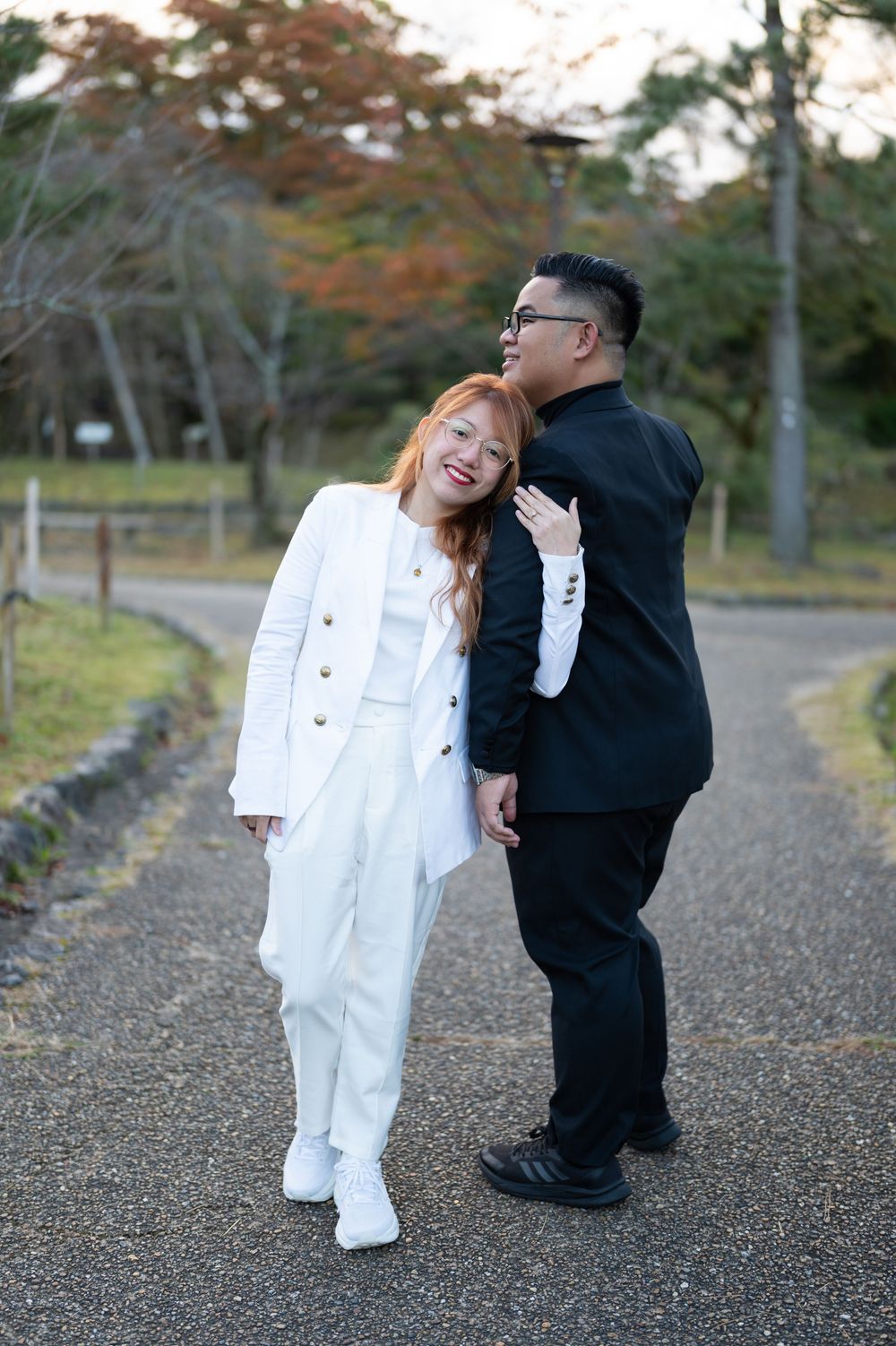 Couple embracing and smiling on tree-lined path surrounded by autumn foliage in Japan.