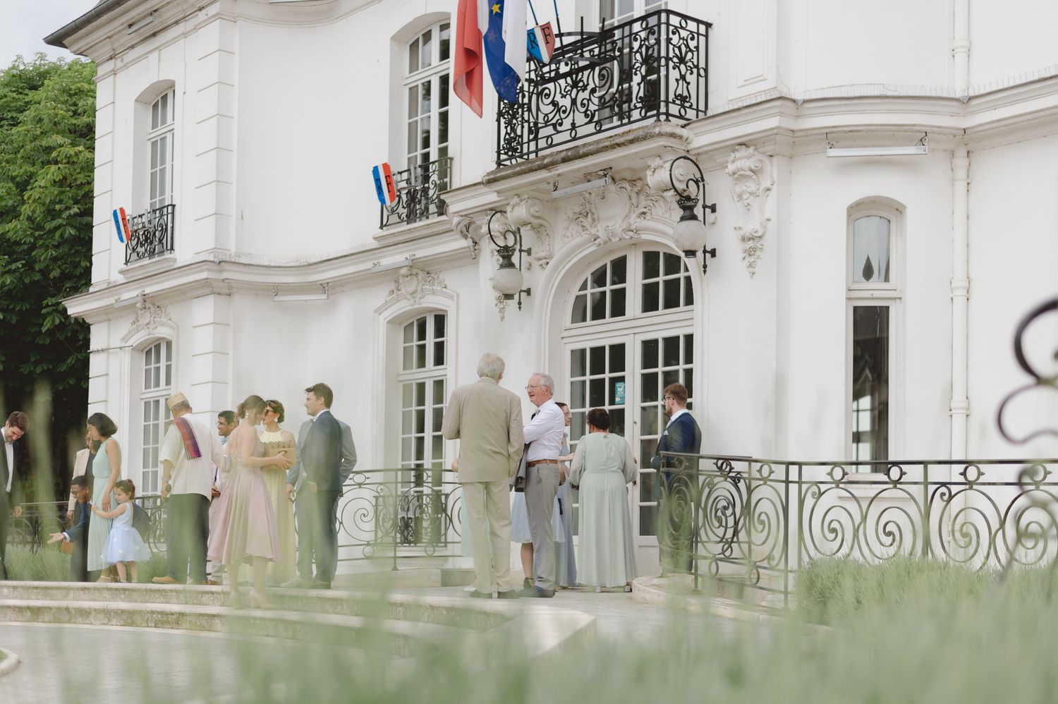 Elegant wedding guests gathering outside a grand white French villa with ornate balcony and national flags.