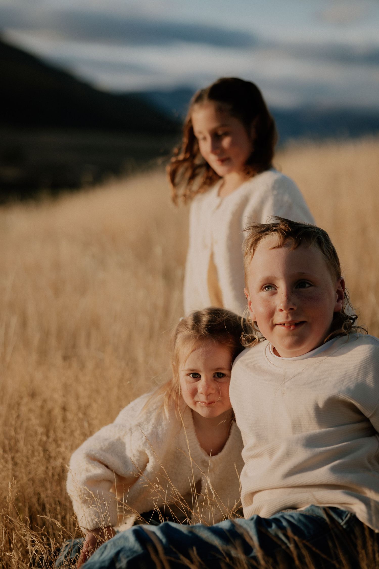 Three children in cream sweaters sit together in a golden wheat field at sunset with mountains in the background.