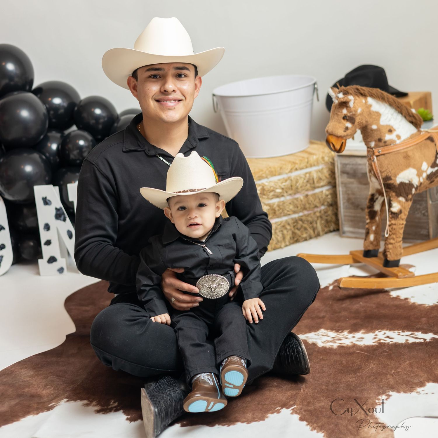A young man and baby in matching black cowboy outfits and white hats pose for a western-themed photo shoot.