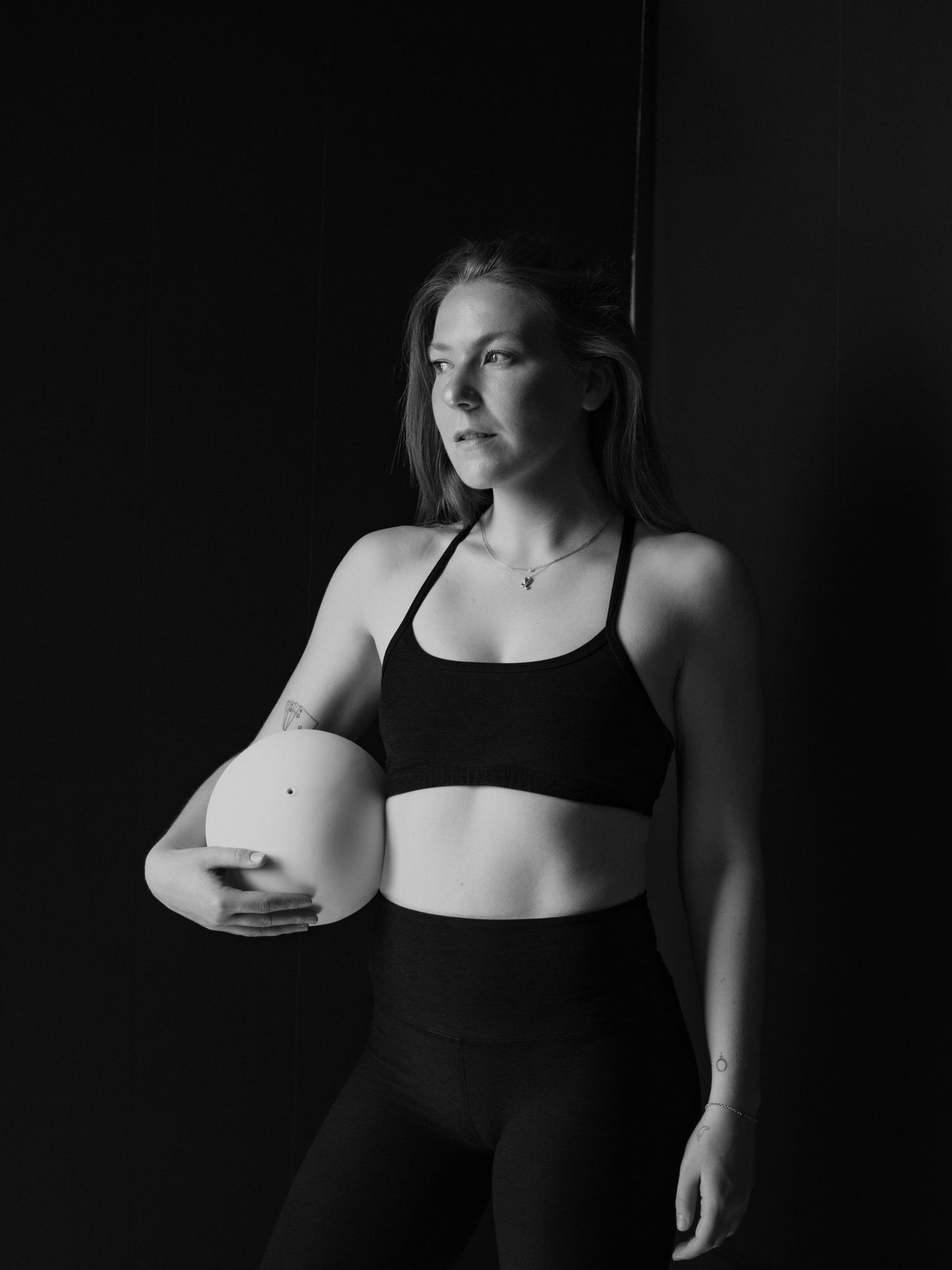 Black and white photo of a fit woman in athletic wear holding a volleyball, posing against a dark background.