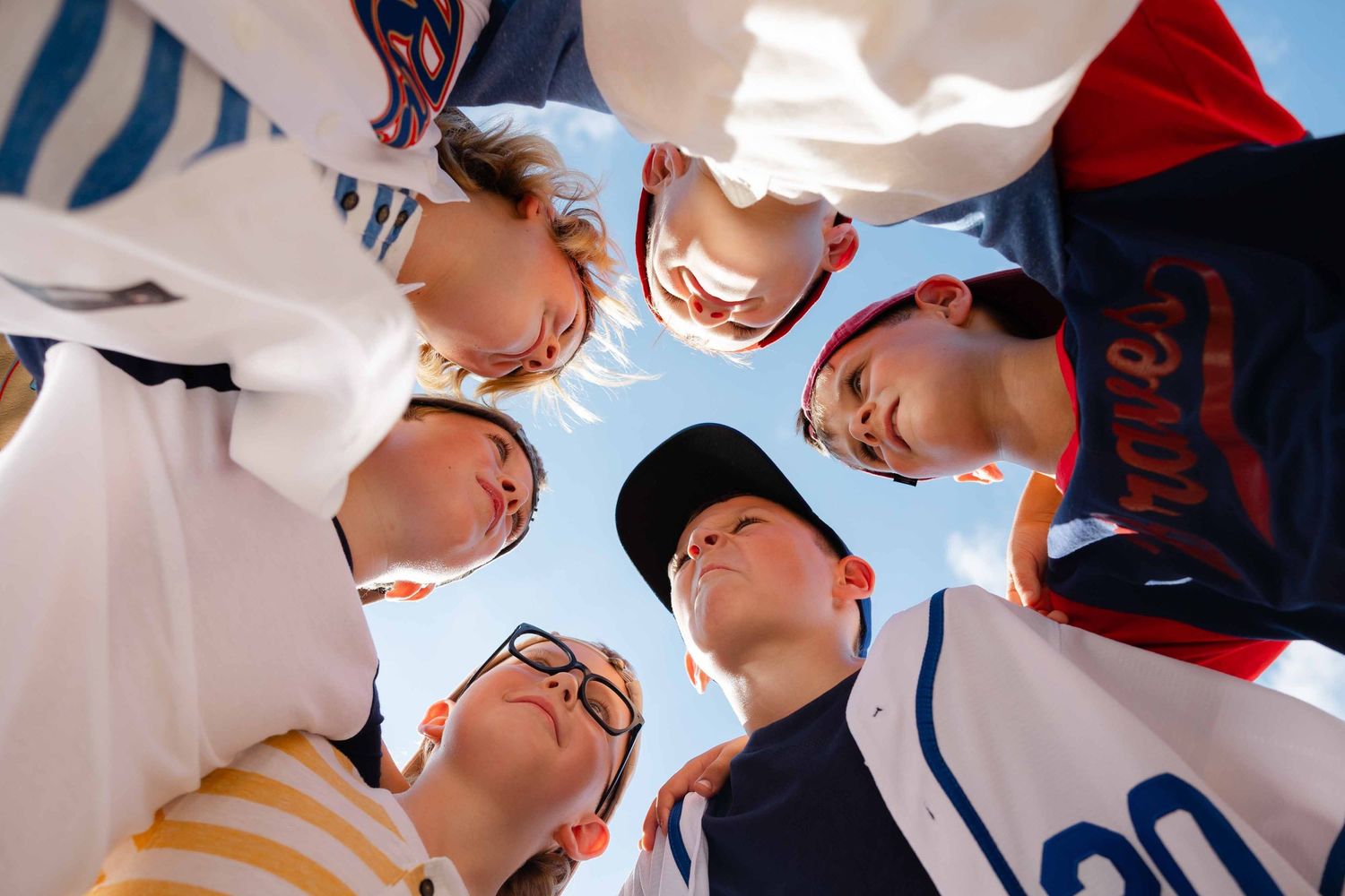 Youth baseball team huddles together in a circle viewed from below against a bright blue sky.