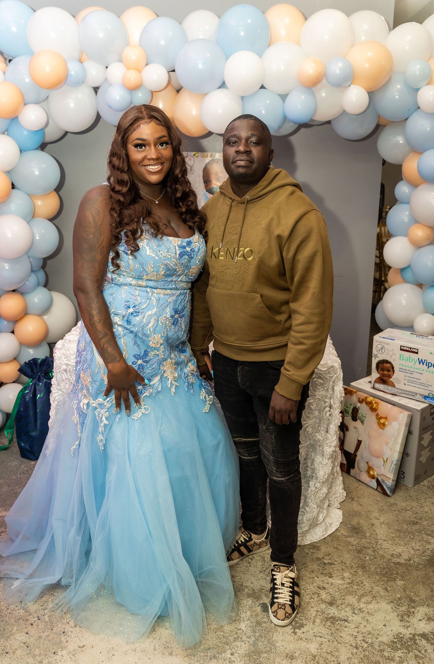 Couple poses together under balloon arch at baby shower, woman in blue floral gown.