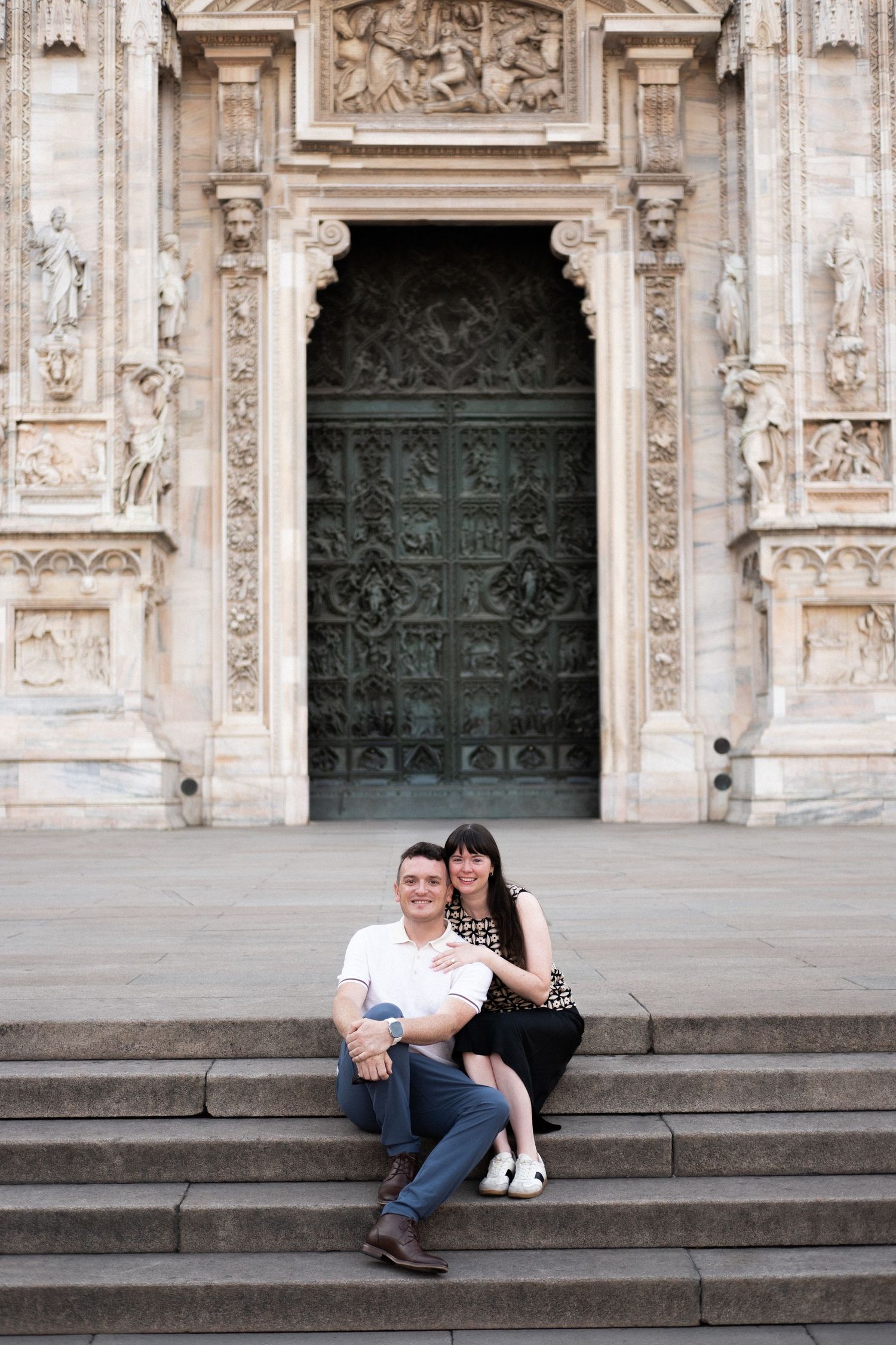 Couple sitting on steps of Milan Cathedral (Duomo di Milano) with ornate bronze doors and marble facade behind them.