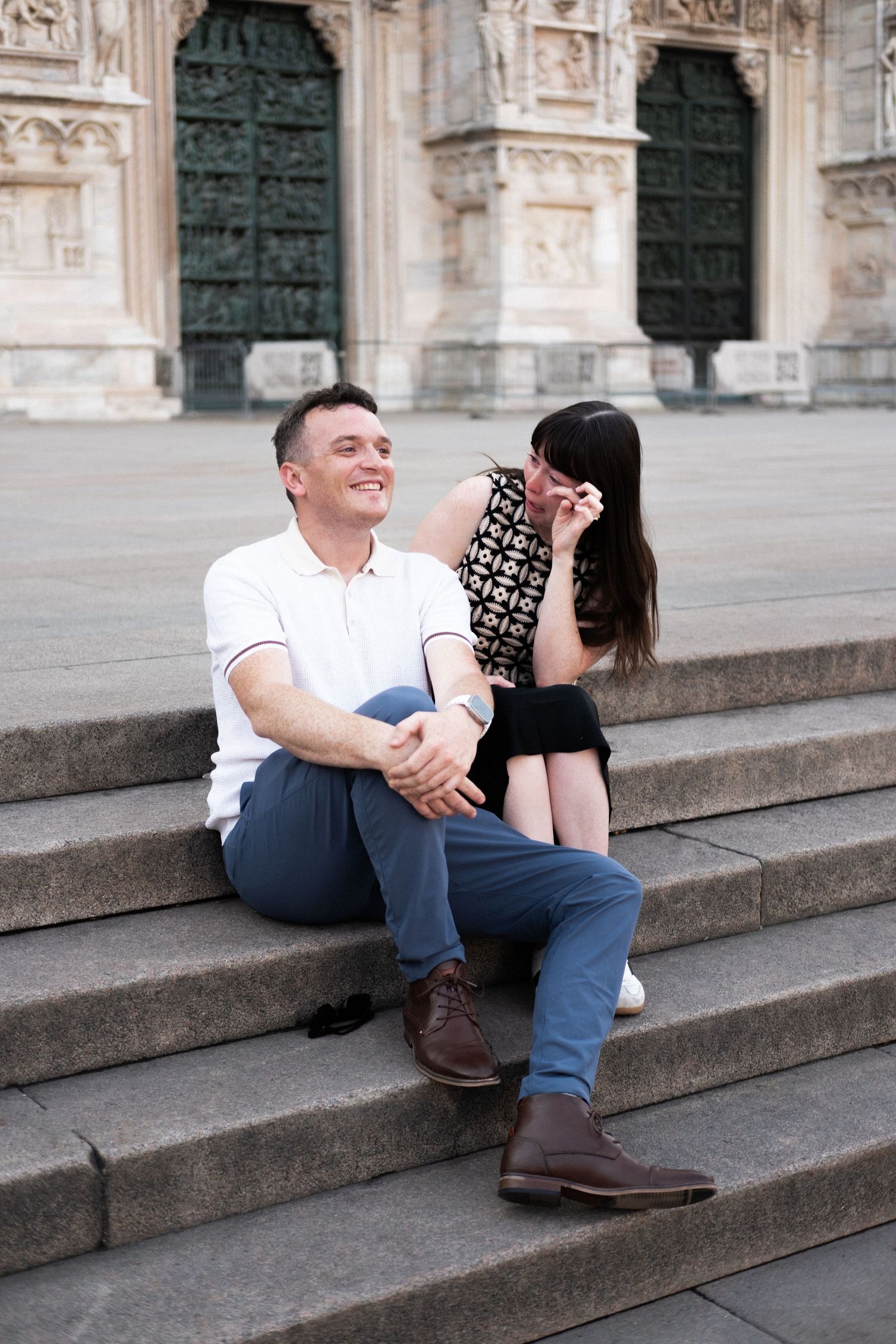 Couple sitting on stone steps outside a historic cathedral, smiling and enjoying a romantic moment together.