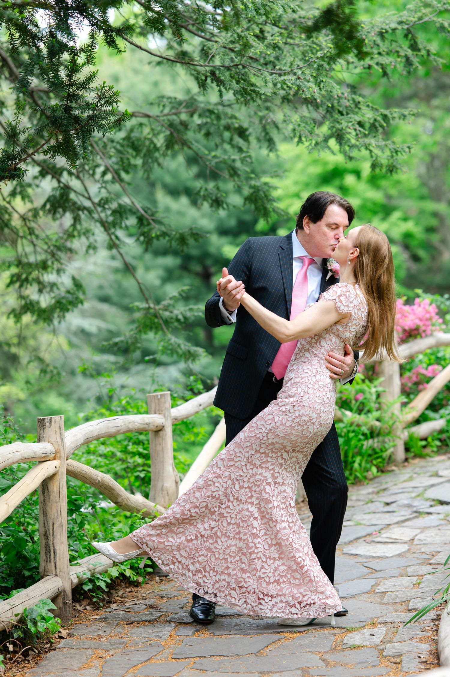 Couple in formal attire walking hand-in-hand on a stone path in a lush green garden, woman holding pink bouquet.