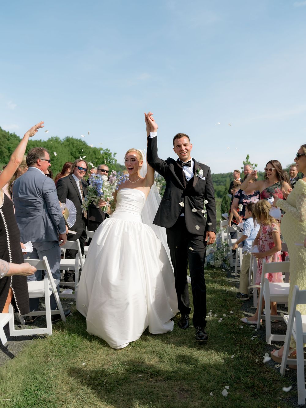 Joyful newlyweds raise hands walking down outdoor wedding aisle as guests toss flower petals.