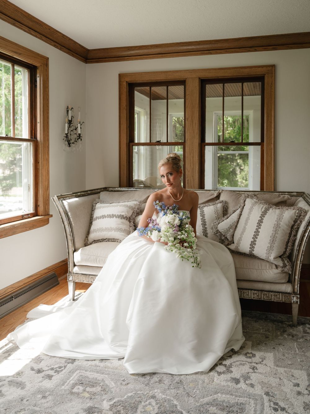 Bride in white ball gown sits on elegant sofa holding floral bouquet in sunlit room with wood-trimmed windows.